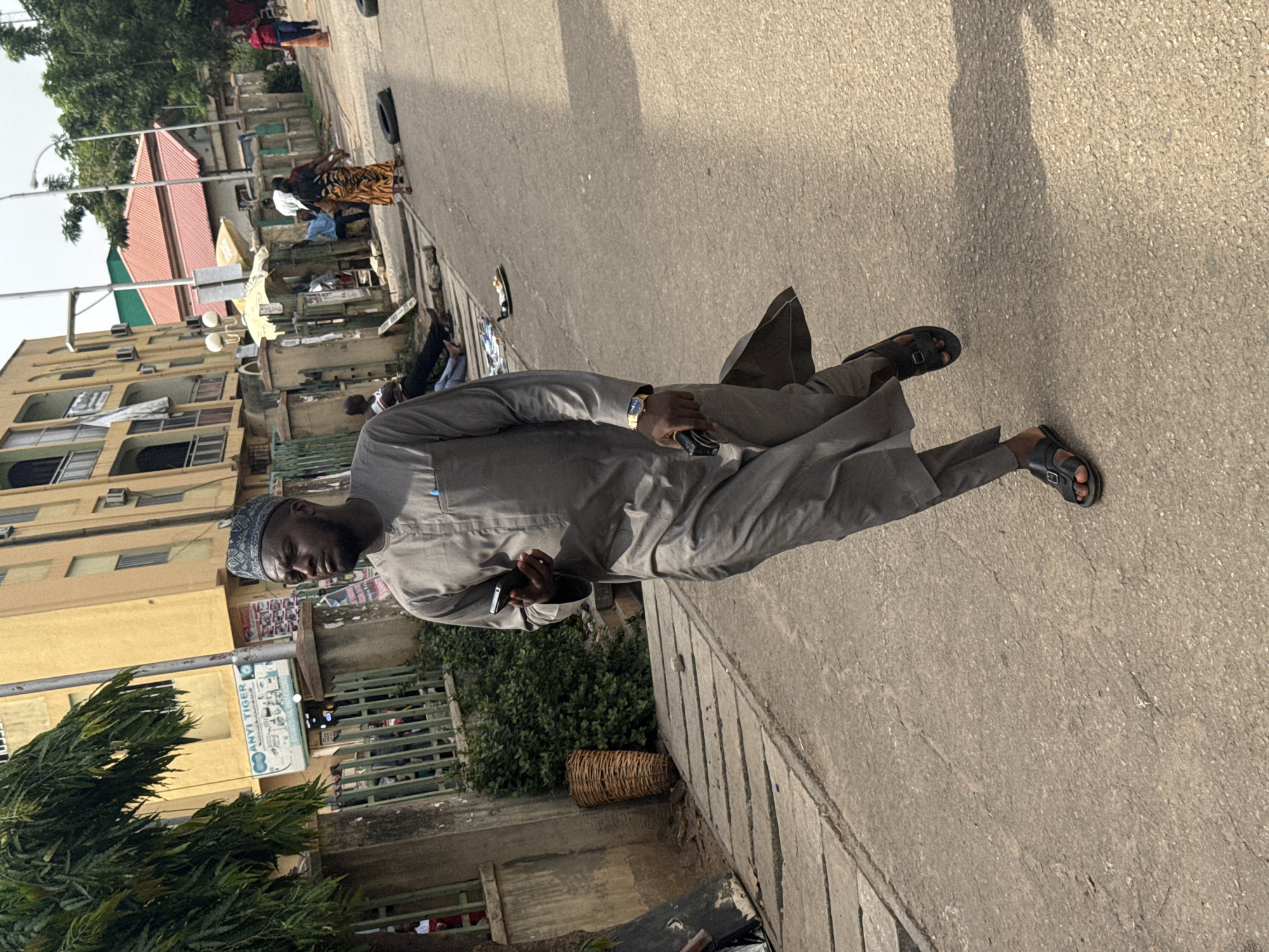 The image, taken in Abuja, Nigeria, depicts a bustling street scene during daylight hours. The primary subject is a man standing in the foreground on a paved sidewalk, oriented towards the viewer. He wears a grey traditional tunic (likely a kaftan or similar attire) and a patterned grey cap. On his left wrist, he has a gold-toned watch, and he holds a dark-colored mobile phone in his right hand. He is wearing dark open-toed sandals. A distinct shadow extends behind him on the ground, suggesting bright sunlight.
Behind the main subject, the street extends into the background, lined with multi-story buildings and various activities. On the left side, a light yellow building with visible air conditioning units stands alongside lush green trees and bushes. Further down the street, on the right side, several people are visible, including one person in a red top and blue jeans, and a group gathered under a white awning or tent. Among this group, one individual wears a distinctive patterned yellow and black skirt or dress, and another is in a white shirt. A dark vehicle tire rests on the paved ground in the middle distance.
The street appears to be a commercial area, with what looks like market stalls or small shops. A utility pole stands prominently, featuring a sign that reads "ANYI TIGER" with smaller, illegible text below, possibly indicating "AUTO SPARE PARTS." Other posters and signs are affixed to walls, but their text is too distant to decipher. A woven basket sits on the ground near the main subject. The sky above appears bright but slightly hazy. The overall atmosphere suggests a typical active urban environment.