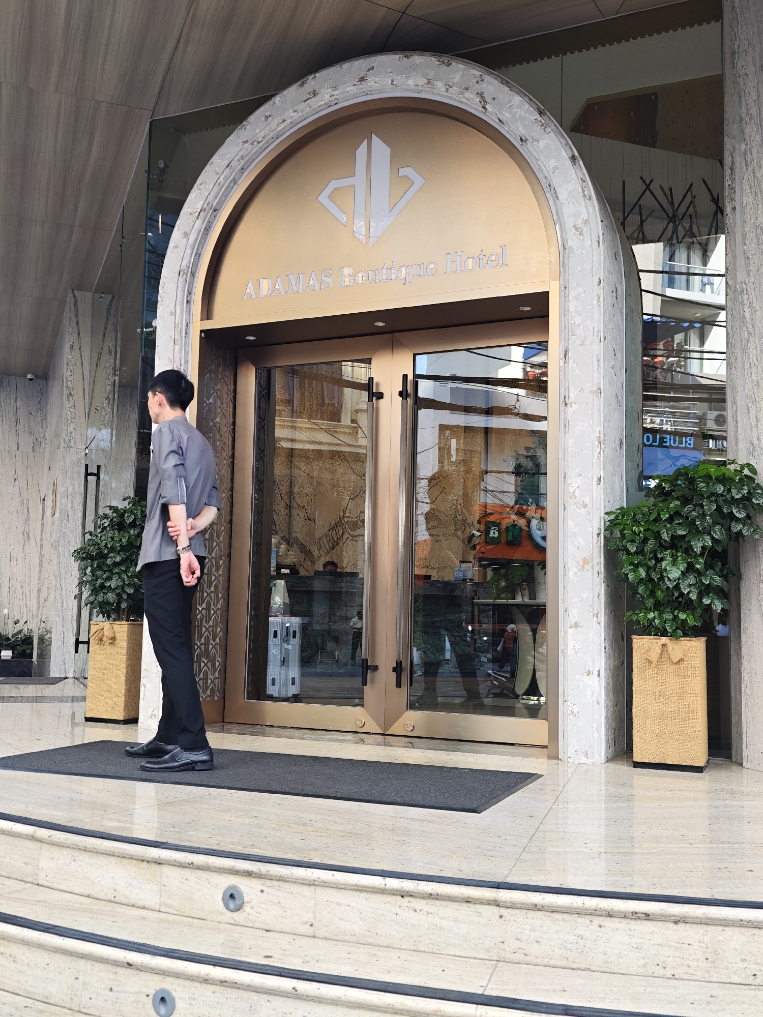 A man in a grey uniform and black pants stands facing away from the camera, with his arms crossed behind his back. He is positioned on a dark grey doormat in front of a large, ornate entrance. The entrance features double glass doors with bronze-colored frames, set within a large, arched stone surround. Above the doors, the words "ADAMAS Boutique Hotel" are visible in silver lettering. To the left of the entrance, a potted plant in a woven basket sits on the floor. To the right, another similar potted plant is visible. The building's facade is made of marble with grey veining. The scene appears to be set during the daytime, with the lighting suggesting fair weather. Reflected in the glass doors are the surrounding buildings and what appears to be an interior space with hanging light fixtures. The overall impression is that of a hotel entrance, with the uniformed individual likely serving as a doorman or concierge. The location context provided is Nha Trang Ward, Vietnam.