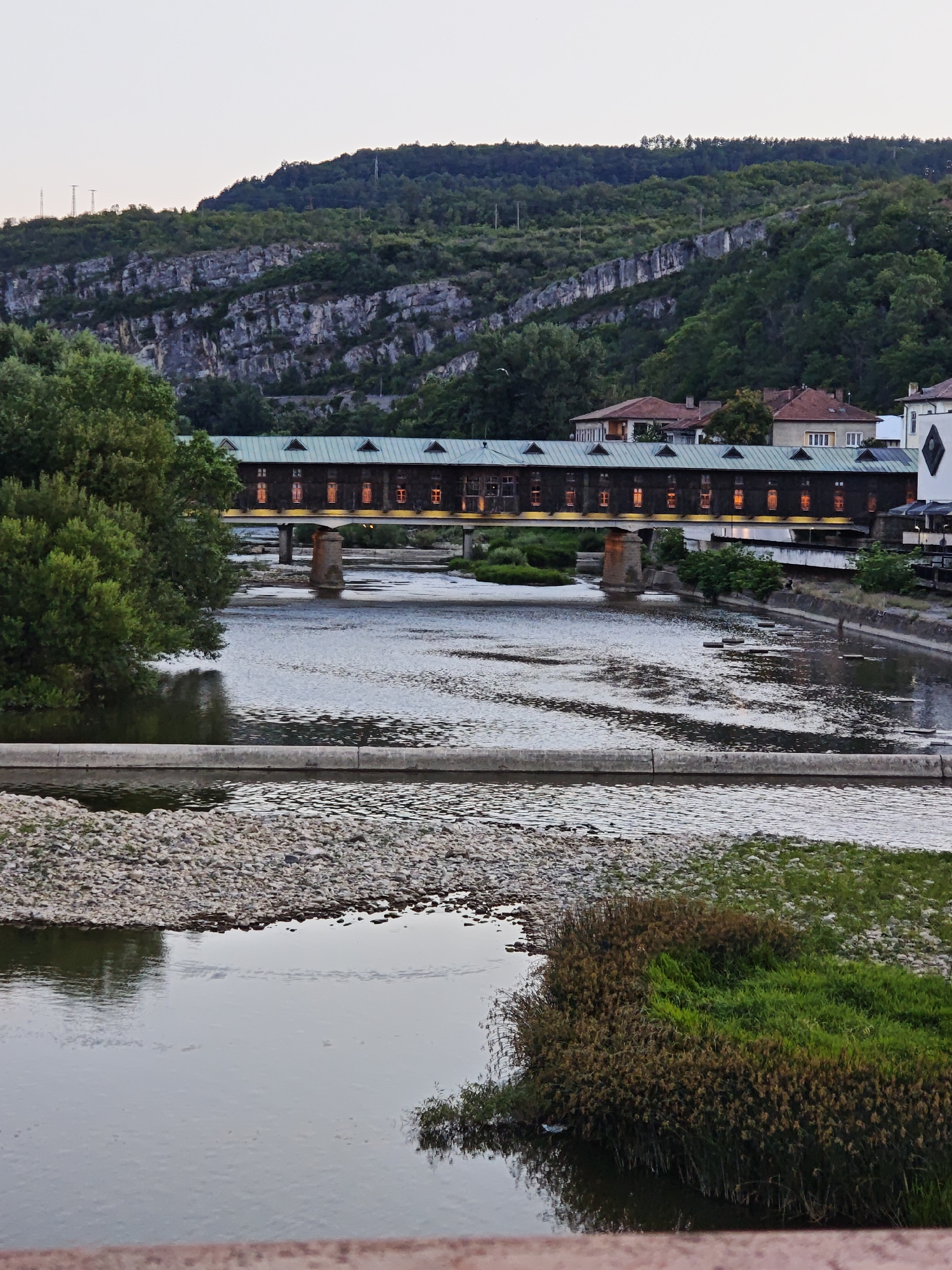 The image depicts the covered bridge over the Yantra River in Lovech, Bulgaria, at dusk. The bridge, a prominent landmark, is illuminated by interior lights visible through its windows, casting a warm glow. It is a long, wooden structure with a green, sloped roof and triangular dormer windows. The river flows beneath the bridge, with a rocky bank on the right side and lush green vegetation on the left. In the background, a forested hill rises steeply, with a rocky cliff face partially visible. Several houses with red tile roofs are situated behind the bridge, indicating the presence of a town. The sky is a pale, dusky hue, suggesting the transition from day to night. There are no people visible in the frame, and the scene is tranquil and serene.