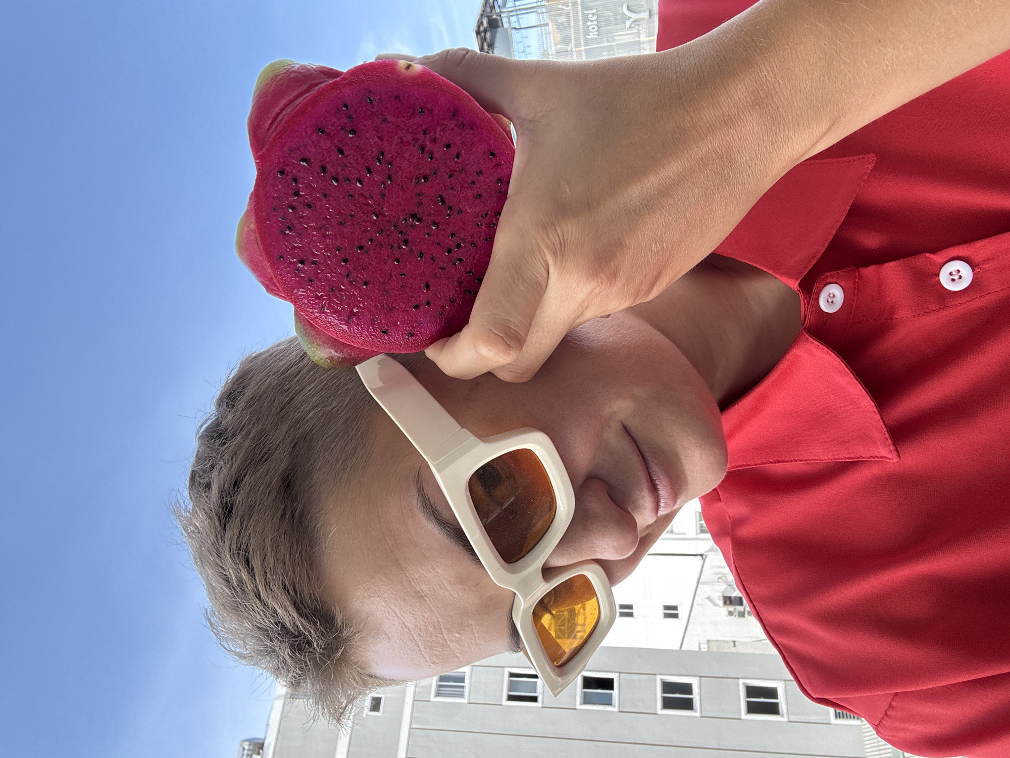 A medium shot captures a person with short, tousled blonde hair, wearing stylish white sunglasses with amber lenses and a vibrant red collared shirt. The person is holding a halved, bright pink dragon fruit to their face, obscuring part of their left cheek. Their gaze is directed downwards, with a slight smile.
In the background, a grey, multi-story building with numerous windows is visible, suggesting an urban environment. Above and beyond the building, a clear blue sky dominates the upper portion of the image. A faint sign, partially obscured, with the word "hotel" in white lettering on a dark background is visible to the right of the building, indicating a hotel in Nha Trang, Vietnam. The overall lighting suggests a bright, sunny day.