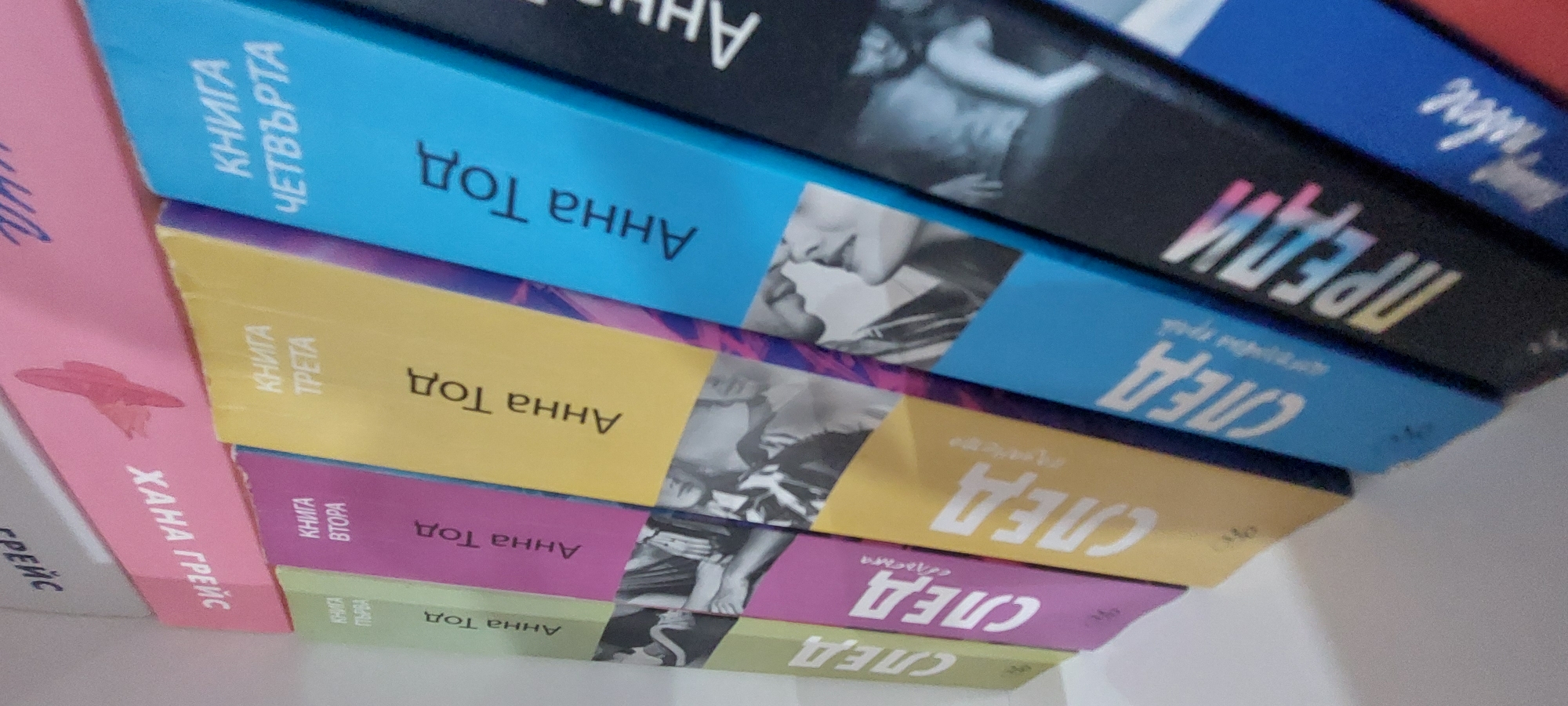 The image displays a series of seven vertically arranged paperback books with one partially visible book, resting on a flat, light-colored surface, presumably a shelf, in a location identified as Yagodovo, Bulgaria. The books are tilted at an angle, presenting their spines to the viewer. All visible text is in Bulgarian Cyrillic. From left to right: 1. **Light green book:** The spine features the title "СЛЕД" (SLED, meaning "AFTER") in large white lettering, with "КНИГА ПЪРВА" (KNIGA PǓRVA, meaning "BOOK ONE") in smaller text below it. The author, "Анна Тод" (ANNA TODD), is printed in black, oriented vertically. A black and white image of a person's torso and arm is visible at the bottom of the spine. 2. **Purple/magenta book:** The spine displays "СЛЕД" (SLED, "AFTER") in white text, with "КНИГА ВТОРА" (KNIGA VTORA, meaning "BOOK TWO") in smaller text. The author is "Анна Тод" (ANNA TODD), printed in black. A black and white image depicting two figures in a close interaction occupies the lower portion of the spine. 3. **Yellow book:** The spine shows "СЛЕД" (SLED, "AFTER") in white text, with "КНИГА ТРЕТА" (KNIGA TRETA, meaning "BOOK THREE") in smaller text. The author, "Анна Тод" (ANNA TODD), is printed in black. A black and white image of a person's upper body is present at the bottom of the spine. 4. **Light blue book:** The spine reads "СЛЕД" (SLED, "AFTER") in white text, with "КНИГА ЧЕТВЪРТА" (KNIGA CHETVǓRTA, meaning "BOOK FOUR") in smaller text. The author is "Анна Тод" (ANNA TODD), printed in black. A black and white image showing a close-up of two faces, seemingly in a tender moment, is at the bottom. 5. **Black book:** The spine displays the title "ПРЕДИ" (PREDI, meaning "BEFORE") in large, iridescent, multi-colored letters. The author, "Анна Тод" (ANNA TODD), is printed in white. A black and white image of a shirtless male embracing a female is visible on the lower spine. 6. **Dark blue book (partially visible):** A small section of the spine is visible, showing partial white Cyrillic text, possibly part of a title like "Любов" (LYUBOV, meaning "Love") or "Вечност" (VECHNOST, meaning "Eternity"). 7. **Pink book (above the light green book, partially visible and inverted):** The spine is pink with an orange butterfly graphic. Inverted Cyrillic text "ХАНА ГРЕЙС" (HANA GREYS, likely "Hannah Grace") is visible, indicating the book is oriented upside down relative to the primary sequence. 8. **White/grey