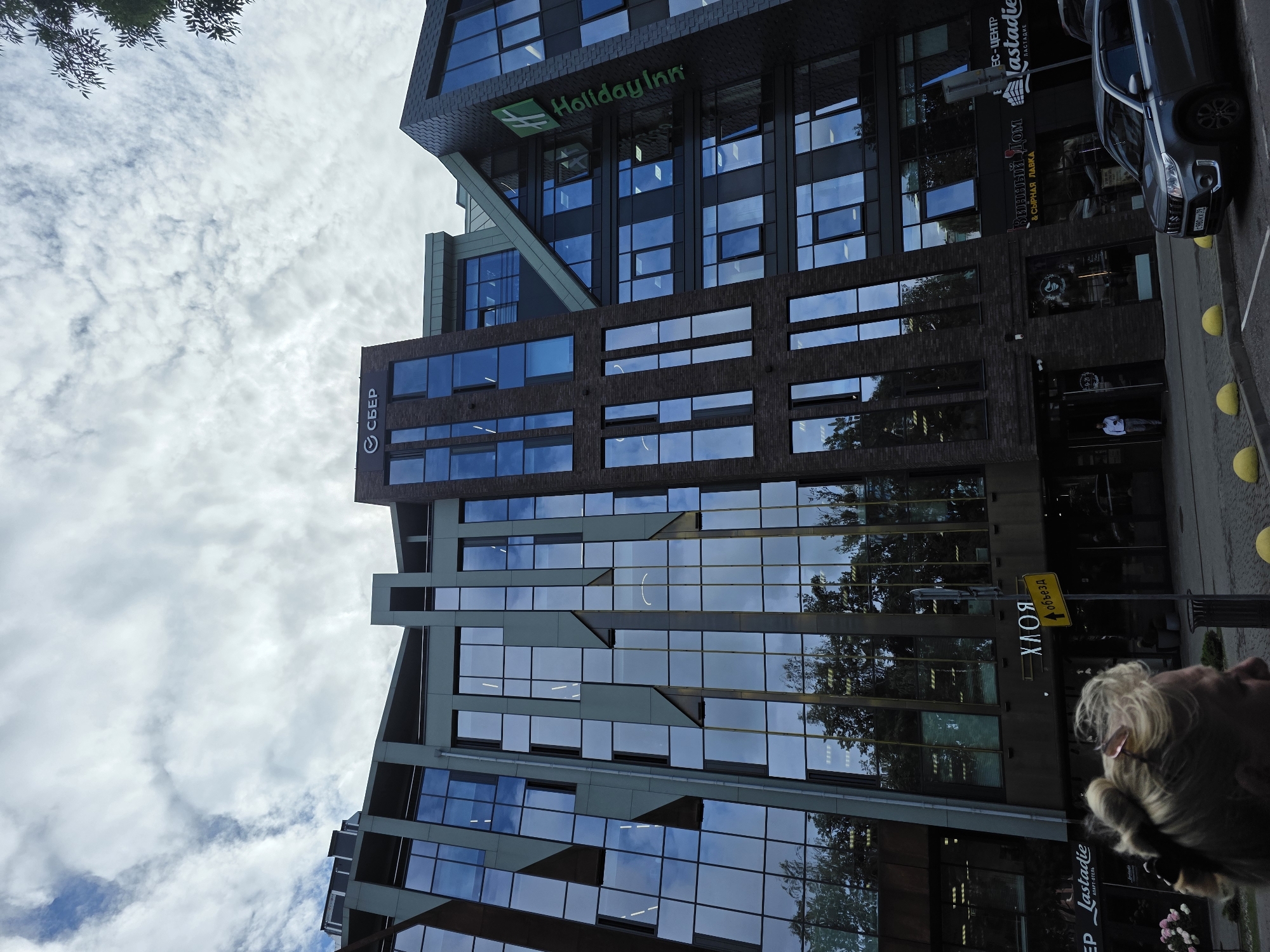 The image captures the facade of a modern multi-story building in Kaliningrad, Russia. The building features a combination of glass and brick elements, with a distinctive angular design. The top of the building displays the logos of "СБЕР" and "Holiday Inn," indicating it houses a Sberbank branch and a Holiday Inn hotel. Below these, the facade has dark-colored windows, reflecting the cloudy sky and surrounding trees. On the ground level, there are storefronts, one with a sign reading "ХЛОЯ," and another with "Lastadie" and "Виный Дом & Сырная Лавка." A yellow sign indicating "объезд" (detour) is visible next to a street lamp. A person's blonde hair and the back of their head are in the foreground, suggesting they are standing or walking near the building. A grey SUV is parked on the street in front of the building. The scene is set on a street with yellow bollards lining the sidewalk.