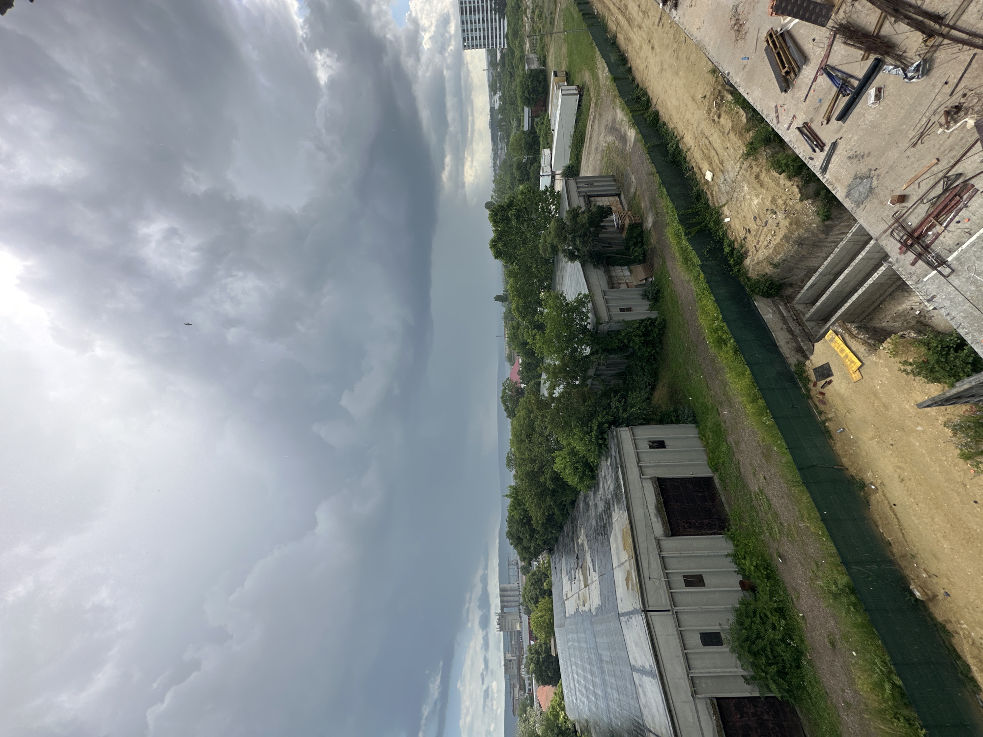The image depicts an outdoor scene, likely during the daytime, with overcast and dramatic cloudy skies, suggesting a storm may be approaching or passing. The perspective is from a high vantage point, looking down and across a landscape that appears to be undergoing construction or development, situated near a vegetated area and a body of water in the distance.
In the foreground, the top of a large, flat-roofed building is visible, with its roof appearing somewhat weathered. To the right, a construction site is evident, with various building materials scattered across a dirt and concrete surface. These materials include rebar, lumber, and what appear to be pre-fabricated concrete elements. A green mesh fence runs along the edge of the construction area, separating it from a grassy strip and more buildings.
Further into the scene, several industrial-style buildings are present, some appearing newer and constructed with metal siding, while others are older and partially overgrown with vegetation. In the middle ground, a line of trees stretches across the landscape, behind which more urban structures, including a tall, modern building with horizontal banding, can be seen. The sky is dominated by dark, heavy clouds, with lighter patches suggesting breaks or the edge of a storm system. A distant, hazy horizon hints at a coastal or large body of water. The overall impression is one of an active development area under a moody, atmospheric sky. No people are visible, and there is no discernible text in the image. The location context of Varna, Bulgaria, implies a European setting.