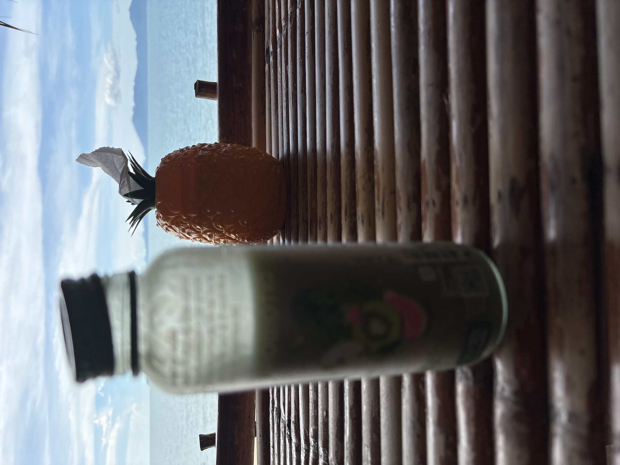 An out-of-focus glass bottle with a black lid and green liquid is in the foreground, slightly to the left of center. To the right of the bottle, a novelty pineapple-shaped object with a crumpled white napkin sticking out of it is in focus. The objects are resting on a horizontally slatted bamboo surface. In the background, a calm, light blue body of water stretches to the horizon, where distant islands or mountains are visible under a blue sky with scattered white clouds. The scene appears to be outdoors, possibly a waterfront cafe or restaurant.