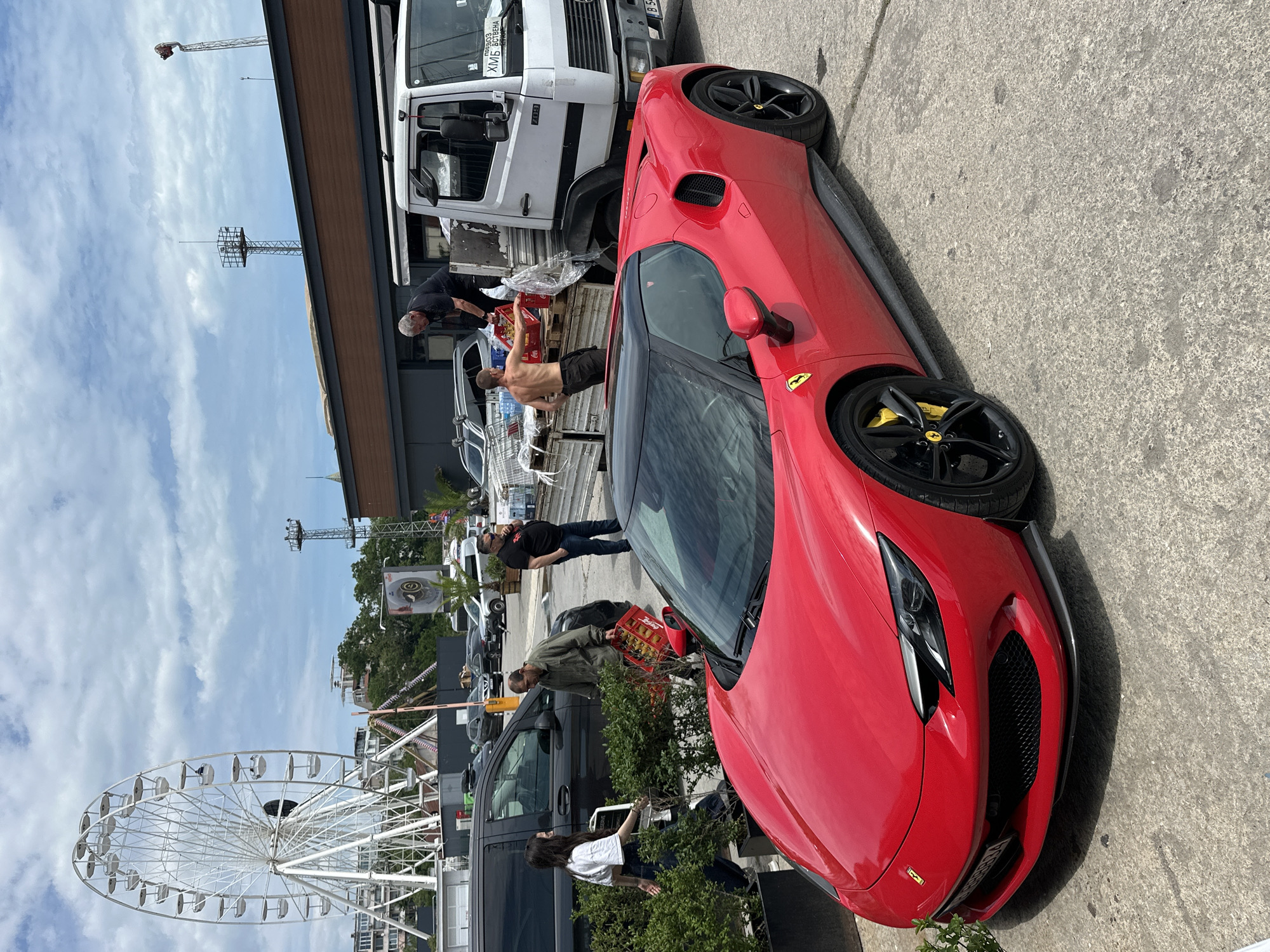 The image depicts a red Ferrari parked on a concrete surface, potentially a parking area. A white truck is parked adjacent to the Ferrari. Several people are visible in the background, some of whom appear to be unloading items from the truck. A Ferris wheel is partially visible in the background. The image was likely taken in Varna, Bulgaria.