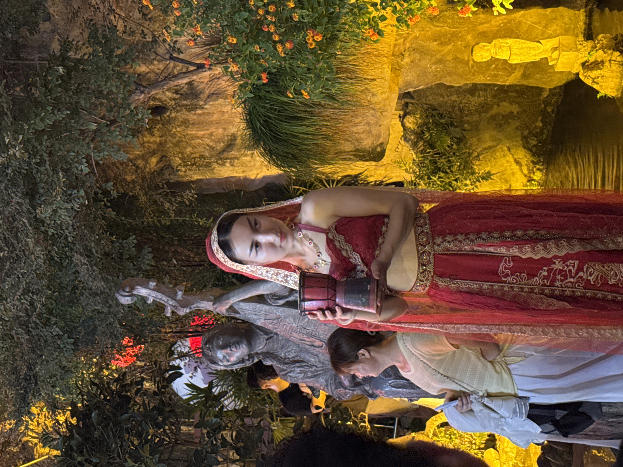 A young woman in a traditional red and gold outfit, complete with a sheer veil and ornate jewelry, holds a small red drum. She stands in front of a statue of a person playing a musical instrument. To her left, a woman in a pale yellow top and white pants holds a white bundle and a small bag. In the background, the scene is illuminated by warm, yellow lighting, highlighting lush greenery, rocks, and a small body of water. A small, light-colored statue of a figure stands on a rock near the water's edge. The overall ambiance suggests an outdoor evening event or display, potentially in Yuzhong District, China, given the context of the query.