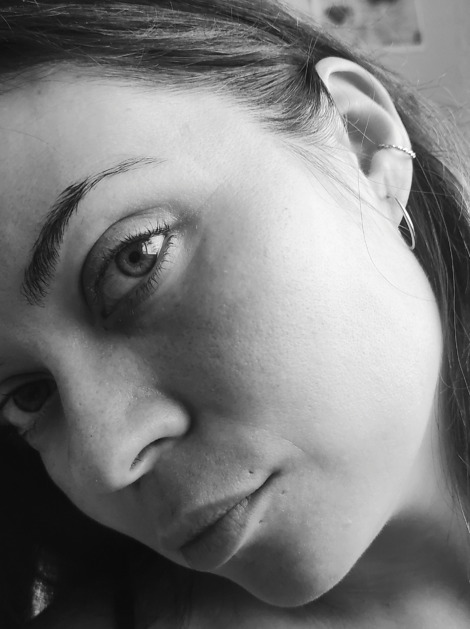 This is a black and white close-up portrait of a person, likely a woman, with her head slightly tilted. The image primarily focuses on the right side of her face (viewer's left), showcasing her eye, eyebrow, nose, mouth, and ear. Dark hair frames her face, with strands visible against her skin and around her ear.
Notable objects include two earrings: a small, textured cuff or ring on the upper cartilage of her ear, and a larger, plain hoop earring hanging from the lower part of the ear. Her right eye is prominently featured, with visible eyelashes, iris, and pupil. A reflection within the pupil suggests an indoor setting with a light source or window. The skin texture is detailed, showing pores and minor imperfections, indicating a realistic portrayal.
The person's expression appears neutral or contemplative, with her eyes looking directly or slightly off-camera. The lighting is soft, creating subtle shadows that enhance the contours of her face. No specific activity is taking place other than posing for the portrait. There is no visible text in the image. The provided location context for this media file is Chaikovsky, Russia.
