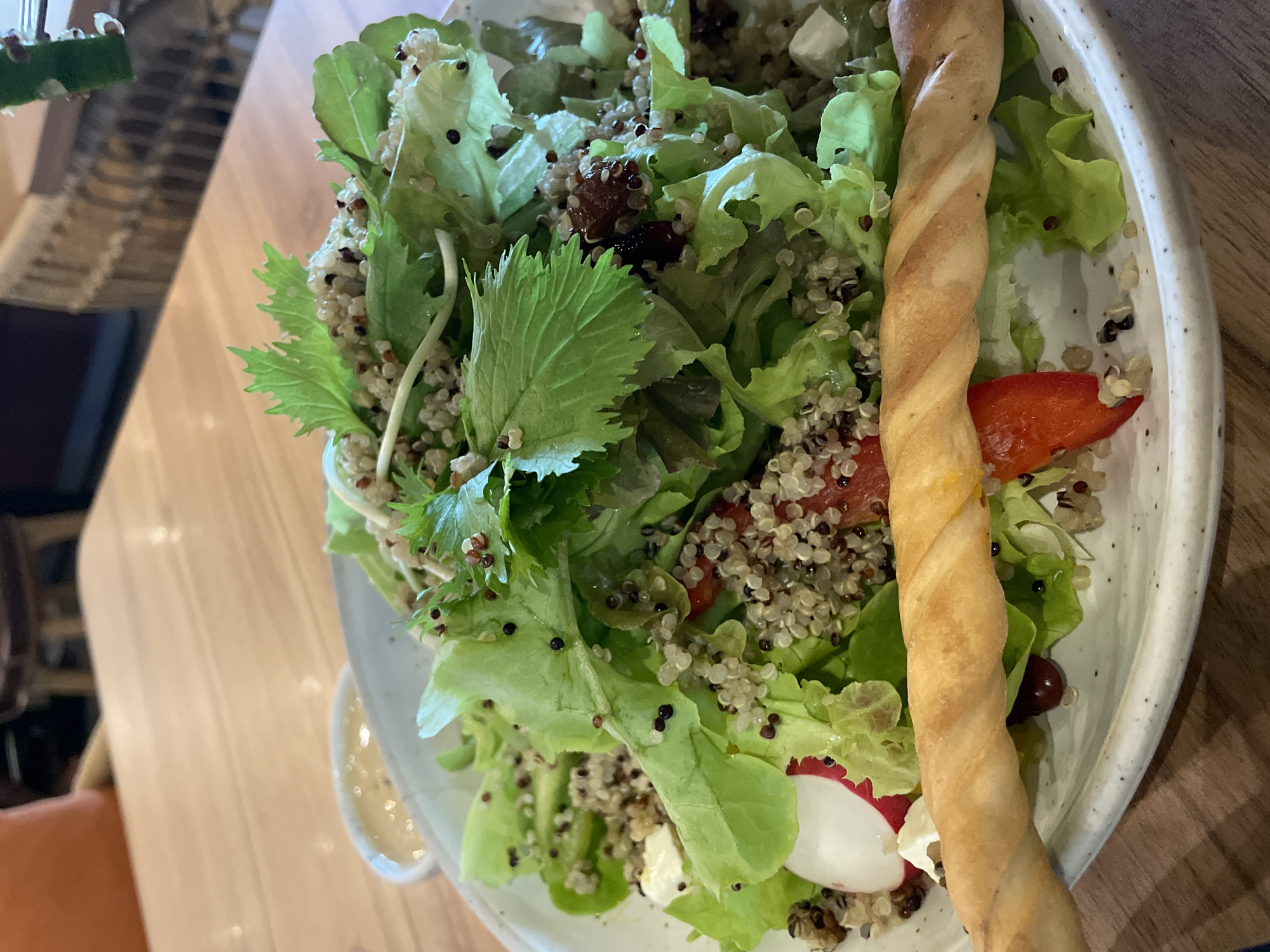 An overhead shot shows a salad on a plate, with a piece of bread in the foreground. The salad contains lettuce, quinoa, feta cheese, radish, and red pepper. The bread is a twisted breadstick. The salad is on a white speckled plate, which is on a wooden table. A small bowl of dressing is visible in the background.