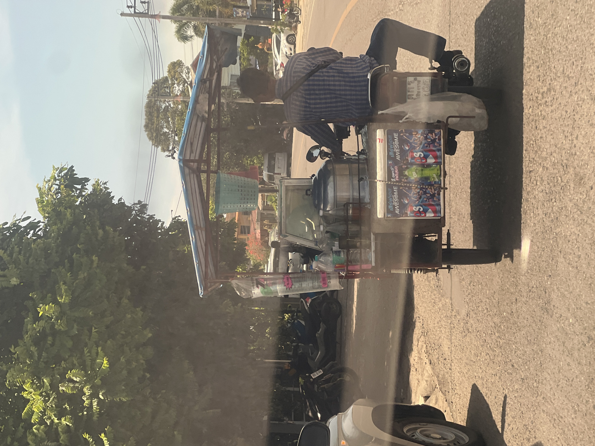 A person rides a motorized cart down a paved road in Ko Samui District, Thailand. The cart is loaded with items including a large metal pot, plastic cups, and several beverage cans. The rider wears a blue and white plaid shirt and dark pants. The cart is covered by a blue tarp. Trees and buildings line the road, and other vehicles are visible in the background. The sky is clear and bright.
