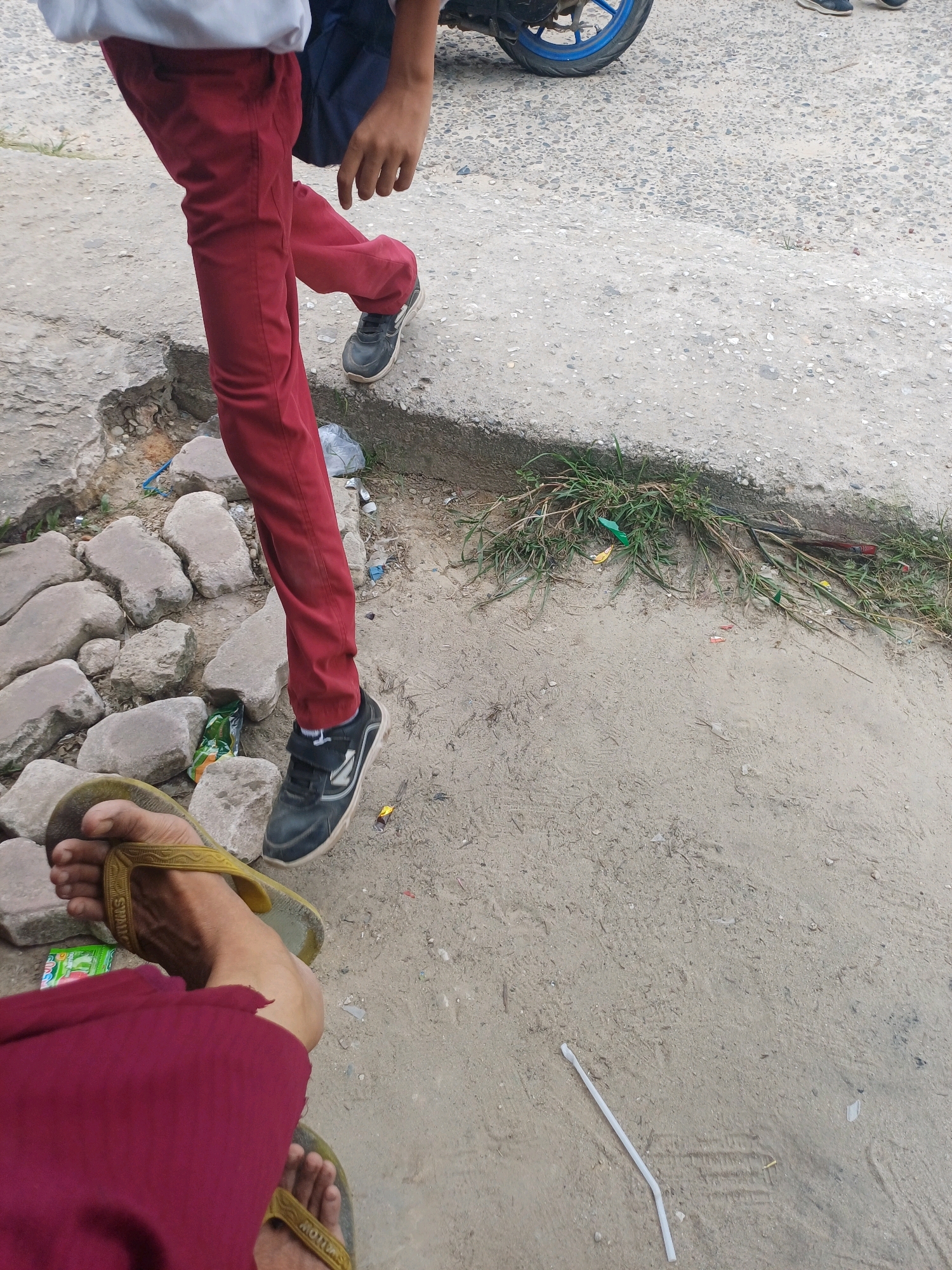 The image shows a person wearing red pants and black sneakers walking on a dirt and concrete pathway. In the foreground, another person's feet are visible wearing yellow flip-flops and a maroon garment. There are rocks and some scattered debris on the ground, including plastic wrappers and a straw. A motorcycle wheel with a blue rim is partially visible in the upper background. The scene appears to be an outdoor urban environment, likely in Dumai City, Indonesia, given the context of the query. The lighting suggests it is daytime.