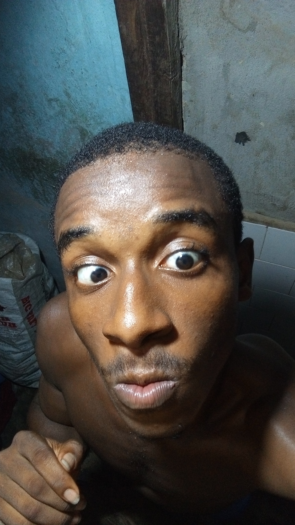 A shirtless man with dark skin is looking directly at the camera with wide eyes and puckered lips. He appears to be indoors, in a dimly lit space with an unfinished ceiling. A large bag is visible in the background to the left of the frame. The city and specific location are unknown.