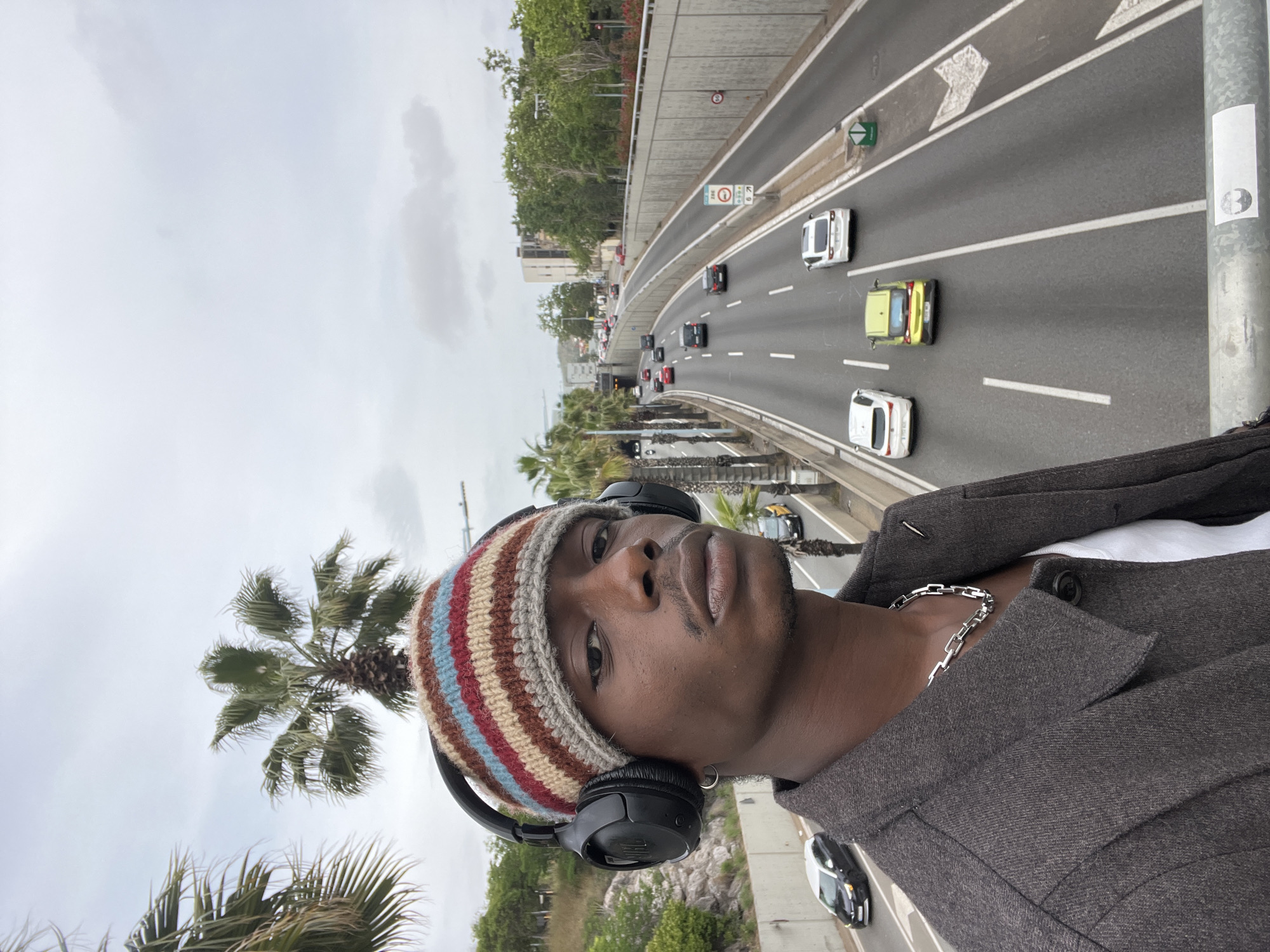 A man is featured in a close-up selfie, positioned on a bridge or overpass with a multi-lane highway stretching into the background. The image is taken during the daytime under an overcast sky. The man, with dark skin, looks directly at the camera with a neutral expression. He wears a multi-colored striped beanie in shades of beige, red, blue, and grey, along with black over-ear headphones. A silver chain is visible around his neck, and he is dressed in a dark grey or brown jacket over a white garment.
The highway below him is busy with several cars in motion, including white, light yellow/green, and darker vehicles. White lane markings and directional arrows are painted on the asphalt. Concrete barriers and lush greenery, notably several palm trees and other tropical foliage, line the sides of the road, indicating a warm climate. Urban buildings are faintly visible in the distance.
Several road signs are identifiable: a circular sign indicating a 50 km/h speed limit, other circular regulatory signs (one with a red border and a diagonal line, another with a red border and a symbol inside), and a green directional arrow sign. A metallic pole in the foreground displays a white rectangular sticker with a dark, circular symbol resembling the Rebel Alliance logo from Star Wars. The scene depicts a lively urban highway environment, consistent with its location in Barcelona, Spain.