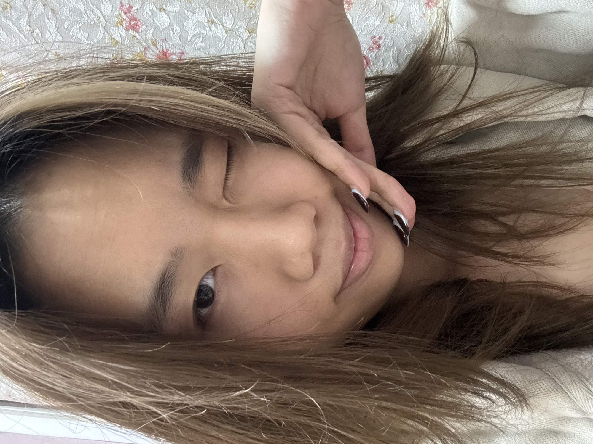 A young woman is lying down indoors, with her head on a patterned surface. She is looking at the camera and winking with one eye. Her hand is near her face, with her fingers close to her cheek. Her hair is light brown. The location is in Shapingba District, China.