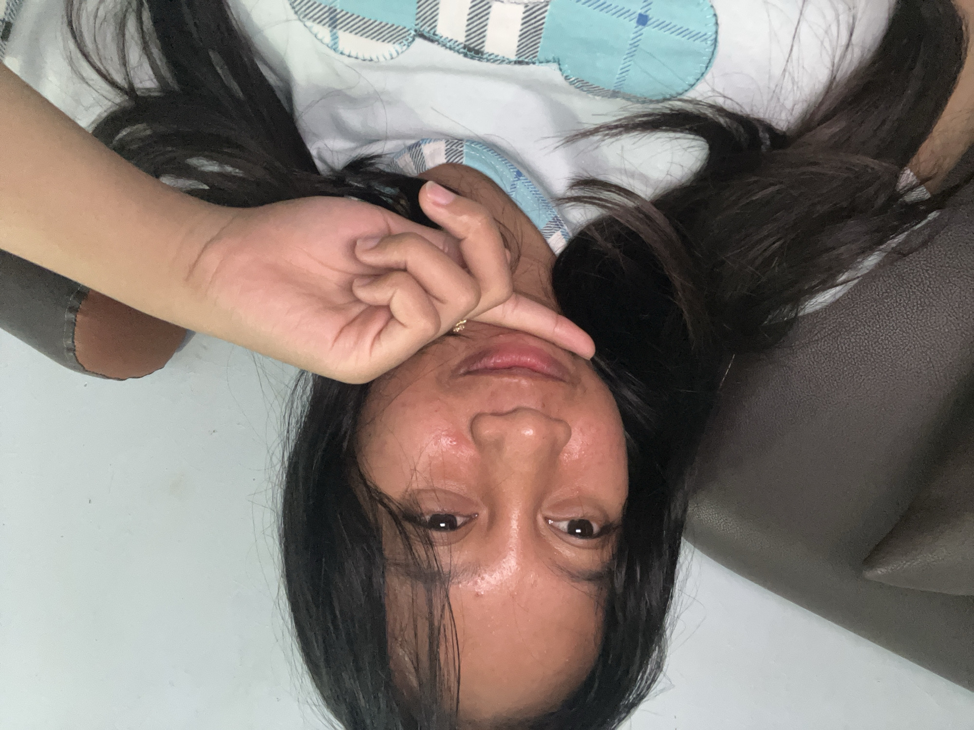 A portrait of a person lying down, with their face close to the camera. The image is rotated 90 degrees clockwise, so the person appears to be upside down. The person has long, dark hair that frames their face. Their skin is visible and appears slightly oily, with some redness on the cheeks. Their eyes are open and looking towards the camera. One hand is raised, with fingers touching the person's face near the nose and mouth. A small gold ring is visible on one of the fingers. The person is wearing a white t-shirt with a blue and white checkered pattern and a blue scalloped trim. The background is a light-colored wall on the left and a dark, textured surface, possibly a couch or chair, on the right. The lighting suggests it might be daytime, but there are no clear external cues to determine the time or weather. The overall emotion conveyed is relaxed or casual, possibly indicating a selfie taken in a home setting. There is no visible text in the image.