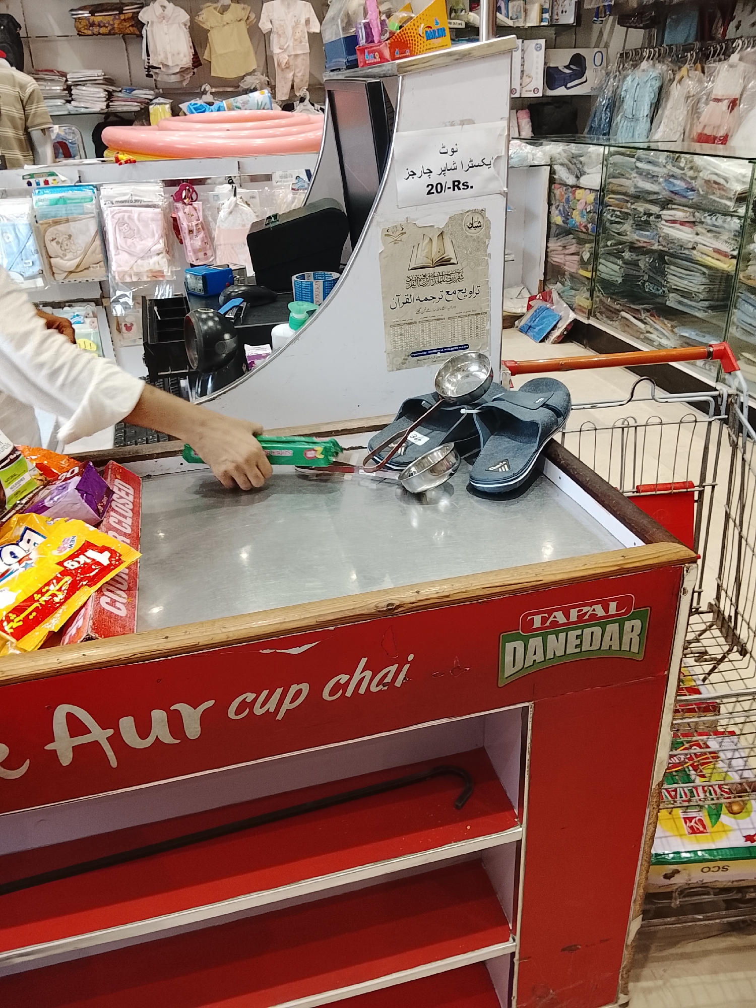 The image shows a cashier's counter in a store, likely in Gulgasht Colony, Pakistan, based on the OCR text. The counter is red and branded with "Tapal Danedar e Aur cup chai". A person is standing behind the counter, handling a package of biscuits. On the counter, there are assorted candies, a pair of sandals, and two metal bowls. A shopping cart is partially visible behind the counter. Shelves beneath the counter are mostly empty, except for a dark, curved metal rod. In the background, shelves are stocked with various goods, including clothing and packaged items. A sign on the counter mentions an extra charge for shopping bags.
