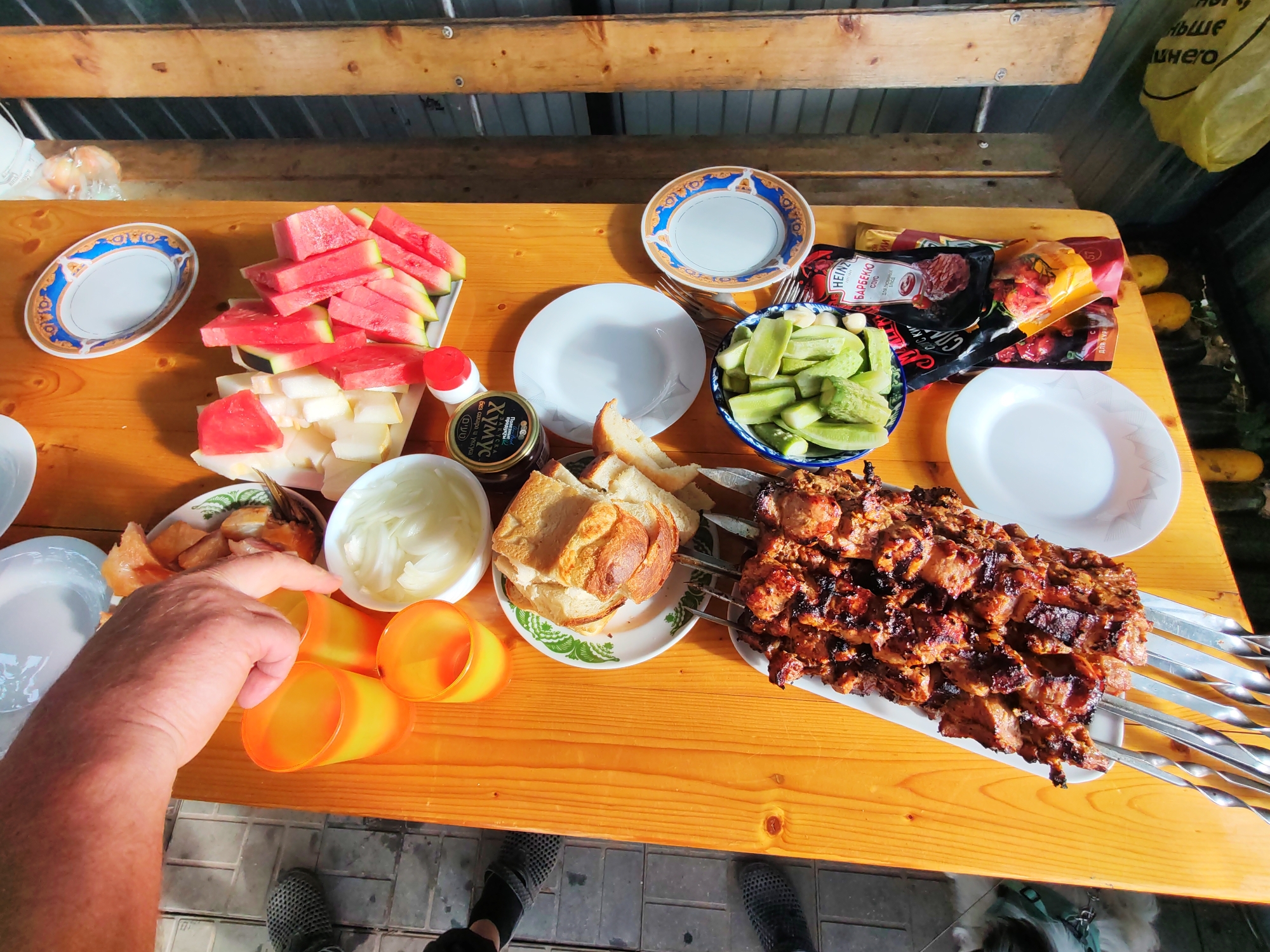 This is a top-down, high-angle, outdoor shot of a wooden table laden with food, set on a paved area that suggests a yard or patio in Pervomaysky, Russia. The lighting indicates it is daytime, likely under a covered structure given the wooden beams above and the diffused light.
The table is covered with various food items. On the left, there's a pile of sliced watermelon and cantaloupe, served on a white platter with green accents. Adjacent to it are three bright orange plastic cups, with a person's hand reaching towards them from the left edge of the frame. Further right, a white bowl contains sliced onions. There is also a plate of sliced bread and what appears to be cooked fish. In the center of the table, a blue and white patterned bowl holds sliced cucumbers. To the right of the cucumbers, several packaged condiments are visible, including Heinz barbecue sauce. A large platter of grilled meat skewers, likely shashlik, dominates the right side of the frame. Several white plates are scattered on the table, some empty. In the background, part of a yellow plastic bag hangs down, and there are some other food items like lemons on the table. The ground beneath the table is paved with grey bricks. The overall scene depicts a casual outdoor meal.