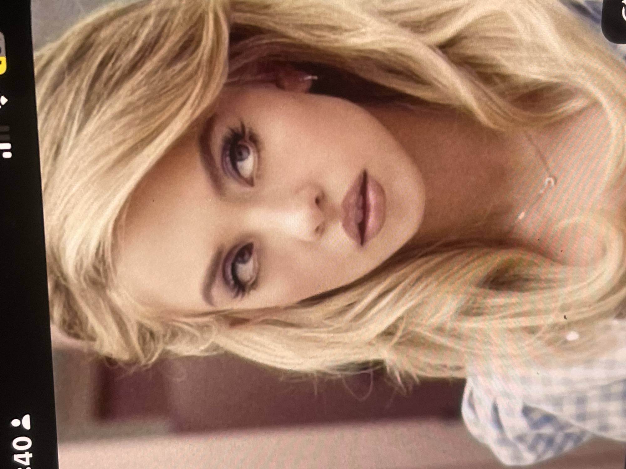 The media file displays a portrait-style photograph of a young woman with long, wavy blonde hair and a fair complexion. Her head is tilted, and she looks directly at the viewer with a direct and slightly contemplative gaze. She has prominent eye makeup, including mascara and eyeliner, and her lips are painted with a nude or pinkish-brown shade. A delicate necklace with a small, light-colored pendant is visible on her neck. Her attire appears to include a light blue and white checkered garment, possibly a shirt, visible on the right side of the frame.
The image itself appears to be a photograph of a digital screen, indicated by the visible screen bezel and interface elements on the left side. These elements include a black bar with white icons, such as a person icon and the number ":40," along with other small white and a yellow icon, suggesting a mobile device interface. The background behind the woman is softly blurred and indistinct, hinting at an indoor setting. There are no specific visual cues within the image to suggest the location of Poltava, Ukraine, but this context is provided separately. The activity depicted is a posed portrait or selfie.