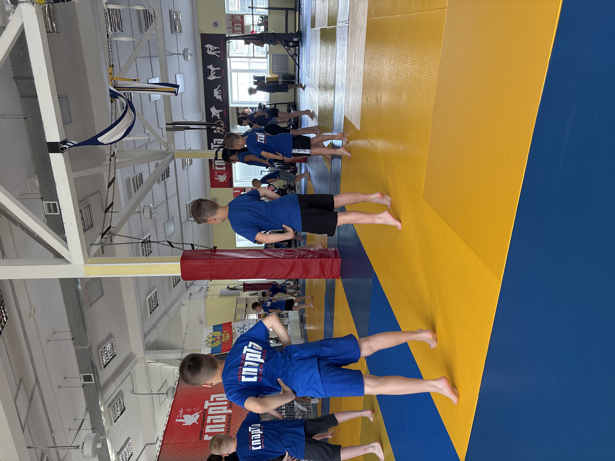 The image depicts a judo training session taking place in a gymnasium. Several young boys and girls, dressed in blue judogi (judo uniforms) and black shorts, are present on a yellow and blue mat. Some are standing in training stances, while others appear to be observing or waiting. The gymnasium features a high ceiling with exposed beams and industrial lighting. Red and white banners with Russian text and symbols are displayed on the walls. In the background, windows reveal daylight, and other gym equipment, including what appears to be a weightlifting station, is visible. The overall atmosphere suggests a sports club or training facility. The location is identified as the Chaikovsky Judo Club in Chaikovsky, Russia.