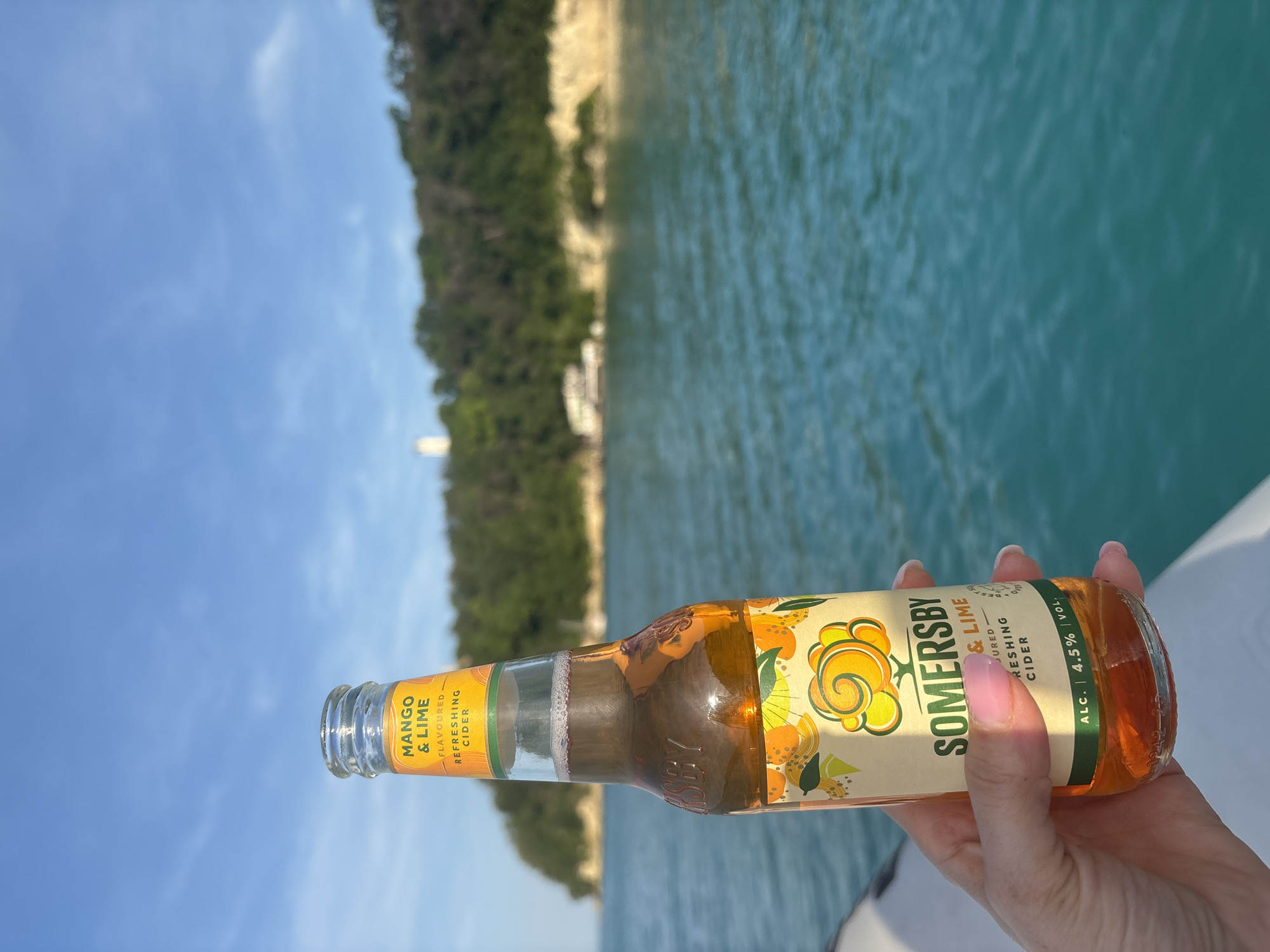 A fair-skinned hand with pink-painted fingernails holds a transparent glass bottle of Somersby Mango & Lime Flavoured Refreshing Cider. The bottle's label is prominent, displaying "SOMERSBY" in green, "MANGO & LIME" in black text on a yellow background, and "FLAVOURED REFRESHING CIDER" below. The label also indicates "ALC. 4.5% VOL". The cider inside the bottle is a translucent amber-orange color with a thin layer of foam at the top. The scene is viewed from a low perspective, with the edge of a white boat or watercraft visible in the lower left foreground.
The midground is dominated by a calm body of turquoise-blue water, showing slight ripples. In the blurred background, a distant, heavily forested landmass is visible. A light-colored, possibly sandy or rocky, cliff face is discernible along the shoreline of the landmass. A prominent white structure, possibly a lighthouse or building, stands on the highest point of the landmass to the left. The sky above is a clear, light blue with minimal, wispy white cloud formations.