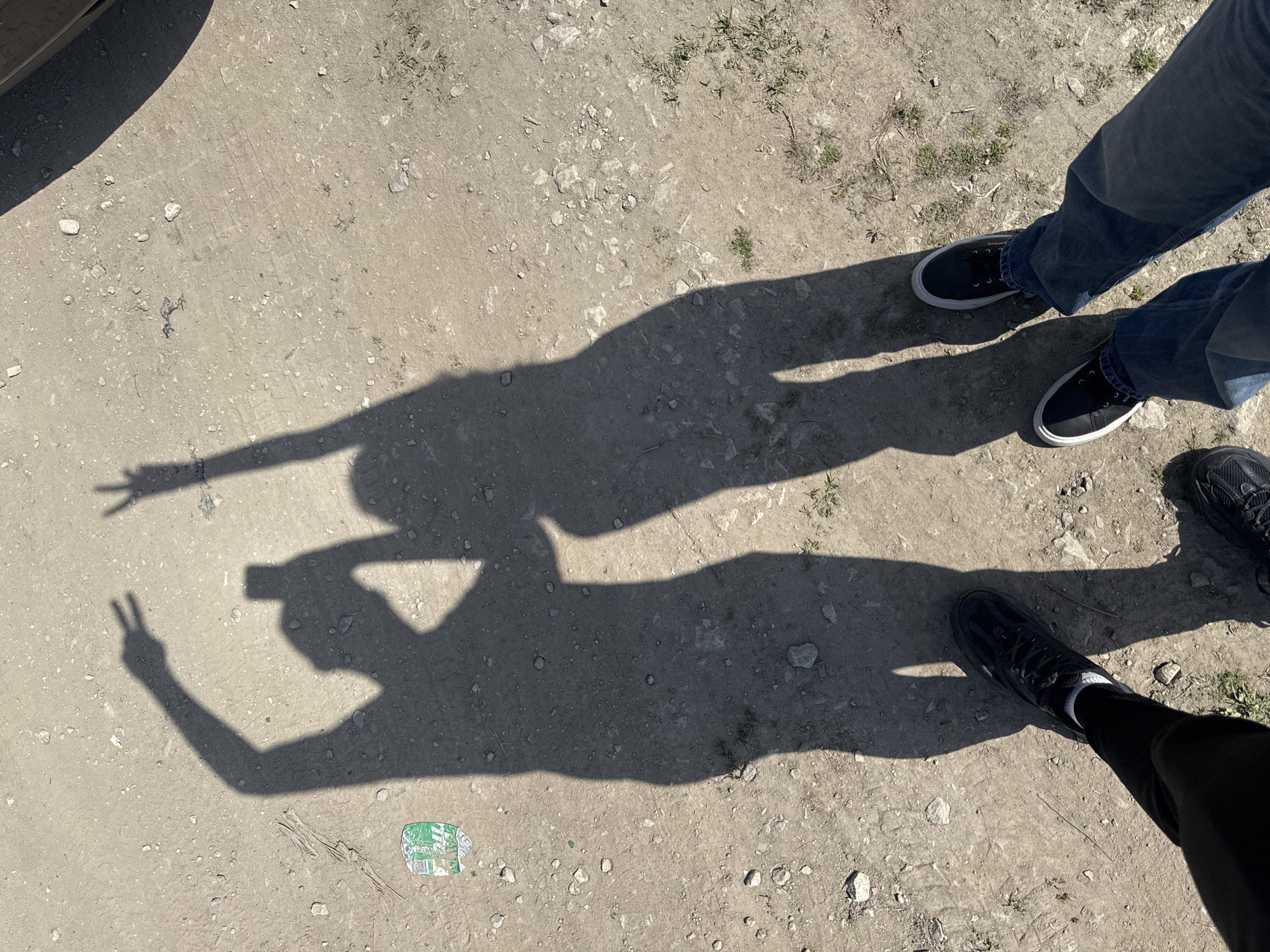 The image depicts an outdoor scene in Boryana, Bulgaria, on a dirt and gravel surface. The shadows of two individuals are visible. One shadow is making a "peace" sign with its fingers, while the other is making a "hang loose" gesture. Visible are the shoes and portions of the legs of both people who are casting the shadows. One individual is wearing blue jeans and black shoes with white soles, while the other is wearing black pants and black athletic shoes with white socks. A crushed and partially obscured beverage container with green, white, and blue text is located on the ground near the "peace" sign shadow. A portion of a vehicle's bumper is visible in the upper-left corner. The overall scene suggests a casual interaction and playful activity between two people.