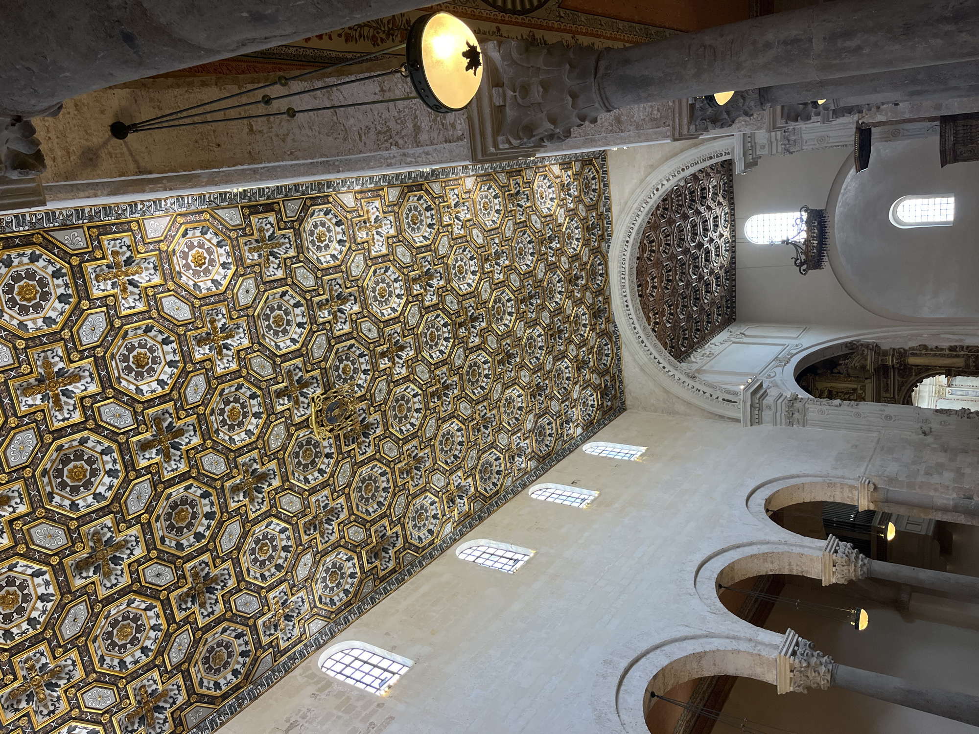 The image shows the interior of a church, possibly the Otranto Cathedral in Otranto, Italy, known for its elaborate mosaic floor. The perspective is angled upwards, focusing on the ceiling and upper portions of the walls. A large, intricately patterned coffered ceiling dominates the view, with geometric designs and possibly religious symbols. The walls are light-colored, featuring arched windows and a series of arches supported by pillars. A large, circular, elaborately carved area is near the top of the frame, possibly around a dome or apse. A hanging light fixture with a simple, round shade is visible. The overall architectural style appears to be Romanesque or Byzantine, reflecting the historical influences of the region. No people are visible in the frame.