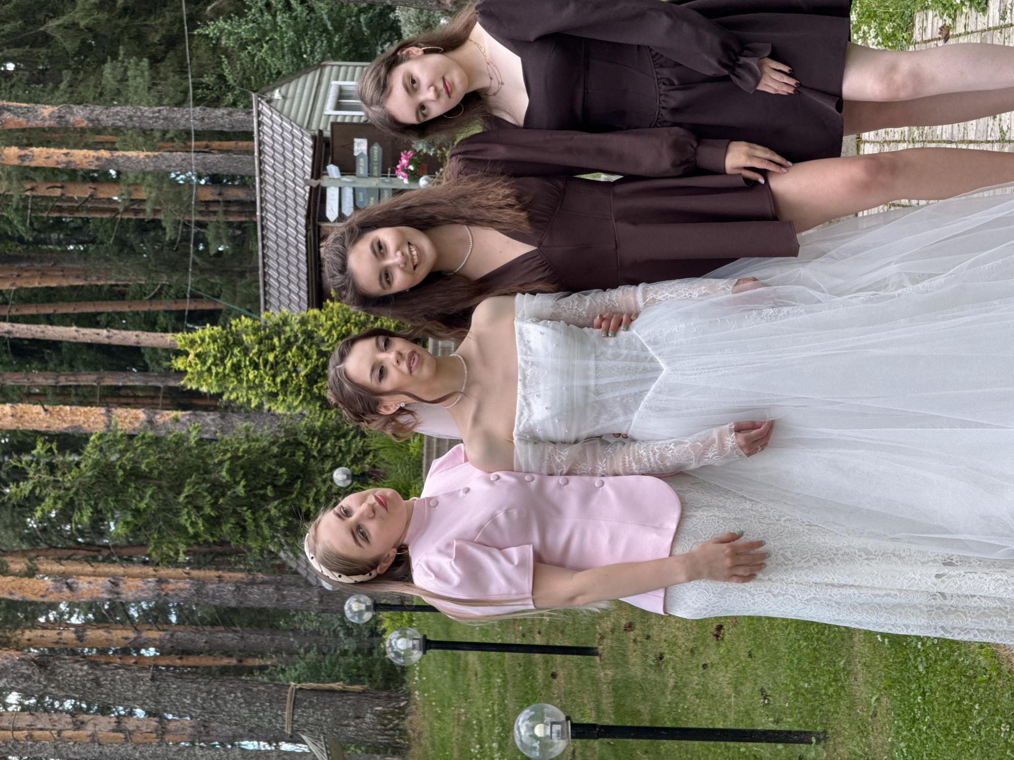 Four young women are posing for a photo outdoors in a grassy area with trees in the background. The woman on the far left is wearing a pink jacket over a white lace skirt. The second woman from the left is wearing a white off-the-shoulder wedding dress with lace sleeves. The third woman from the left is wearing a short brown dress. The woman on the far right is wearing a dark brown dress with puff sleeves. They are all looking at the camera and smiling. In the background, there is a small wooden building with a gray roof, several trees, and some outdoor lights. The scene appears to be a celebration or event, possibly a wedding given the presence of the wedding dress. The overall setting suggests a rural or suburban location, typical of the outskirts of Votkinsk, Russia, known for its natural beauty and dacha culture. The lighting indicates it is daytime, with soft natural light.