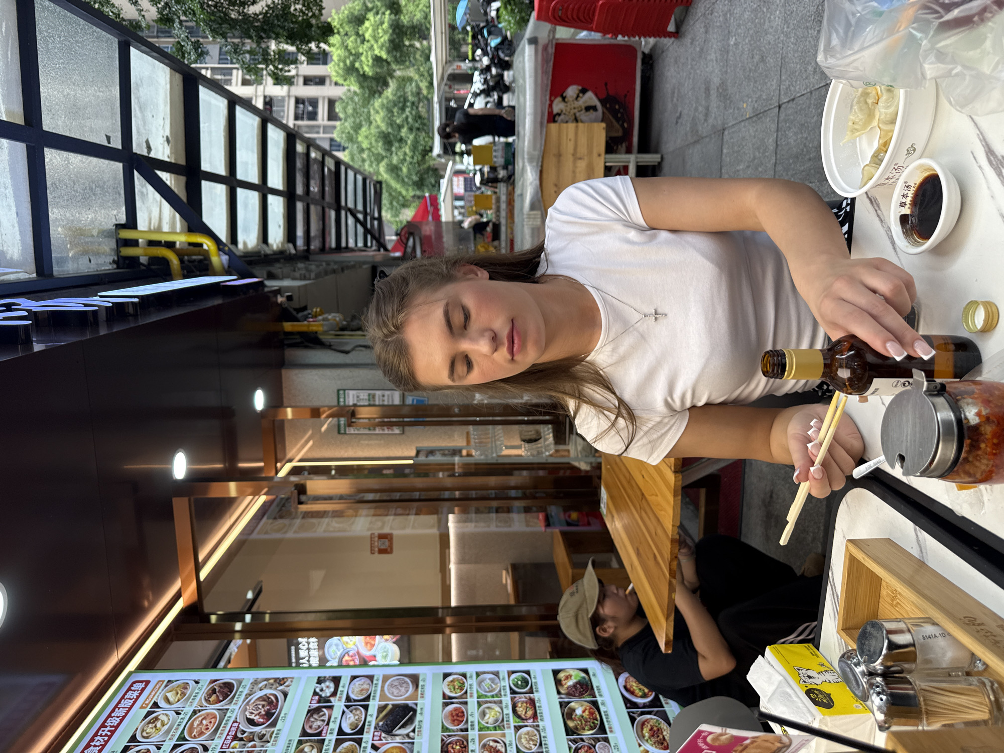 In Haizhu District, China, a young Caucasian woman with blonde hair pulled back is seated at a white table outdoors, facing slightly to her left, with her eyes closed. She is wearing a white t-shirt and a silver cross necklace. Her hands are positioned over the table, with chopsticks held in her right hand, appearing to interact with food. A brown glass bottle with a gold cap is placed next to her right hand. In front of her is a small white dish with dark liquid, likely a dipping sauce. A white bowl containing several dumplings is to her right. A plastic bag is also visible on the table. The table is cluttered with various items including a wooden tray with two salt and pepper shakers and a small wooden container, a box of yellow tissues, and a jar of red chili paste. In the background, another person wearing a black shirt and a tan baseball cap is visible, seated and smoking, with a wooden table between them. The setting appears to be a street-side dining area with a covered walkway overhead supported by metal beams and a corrugated roof. Buildings and trees are visible in the distance, along with parked motorcycles. A large menu board with Chinese text and images of food items is mounted on the wall of a building to the left.