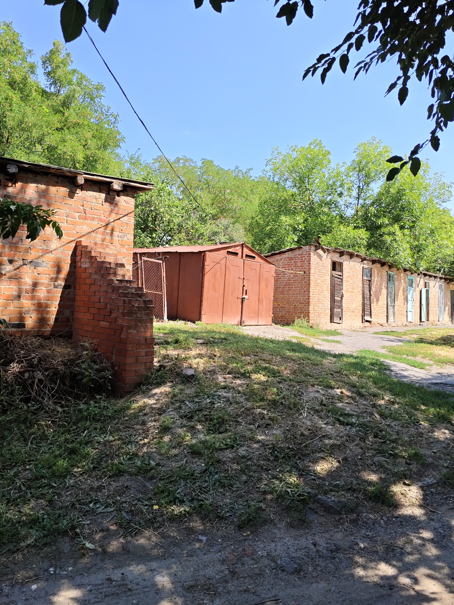 This image depicts a row of old, weathered brick garages situated in Pukhlyakovskiy, Russia. The garages are a mix of brick and corrugated metal construction, with some showing signs of disrepair. The foreground is dominated by a grassy, uneven slope leading up to the garages, with some debris and dry vegetation visible. In the background, lush green trees provide a natural backdrop against a clear blue sky. The overall impression is one of an aged, utilitarian area, likely a communal garage space. There are no people or visible actions taking place in the scene.