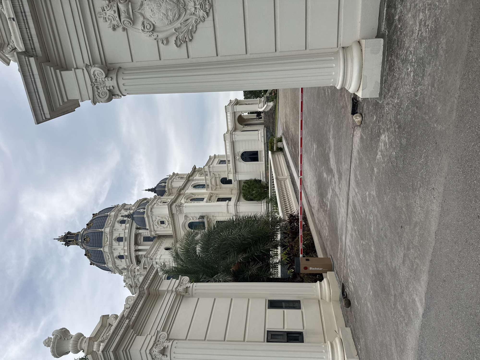 The image depicts the entrance to a grand white building with a large blue dome, situated in Hoa Lư Ward, Vietnam. The building features classical architectural elements such as columns, ornate carvings, and multiple smaller domes. In the foreground, a grey asphalt road leads to the entrance, which is partially blocked by a red and white retractable barrier. A brown automated barrier gate is also visible, with the brand "Blackstar" printed on it. Two large white pillars frame the right side of the entrance, with decorative carvings on the wall behind them. On the left side, another white structure with a pillar and a small dark window is present. A palm tree and other lush greenery are visible behind the barrier, leading towards the main building. The sky is overcast, suggesting a cloudy day. No individuals are clearly discernible in the scene.