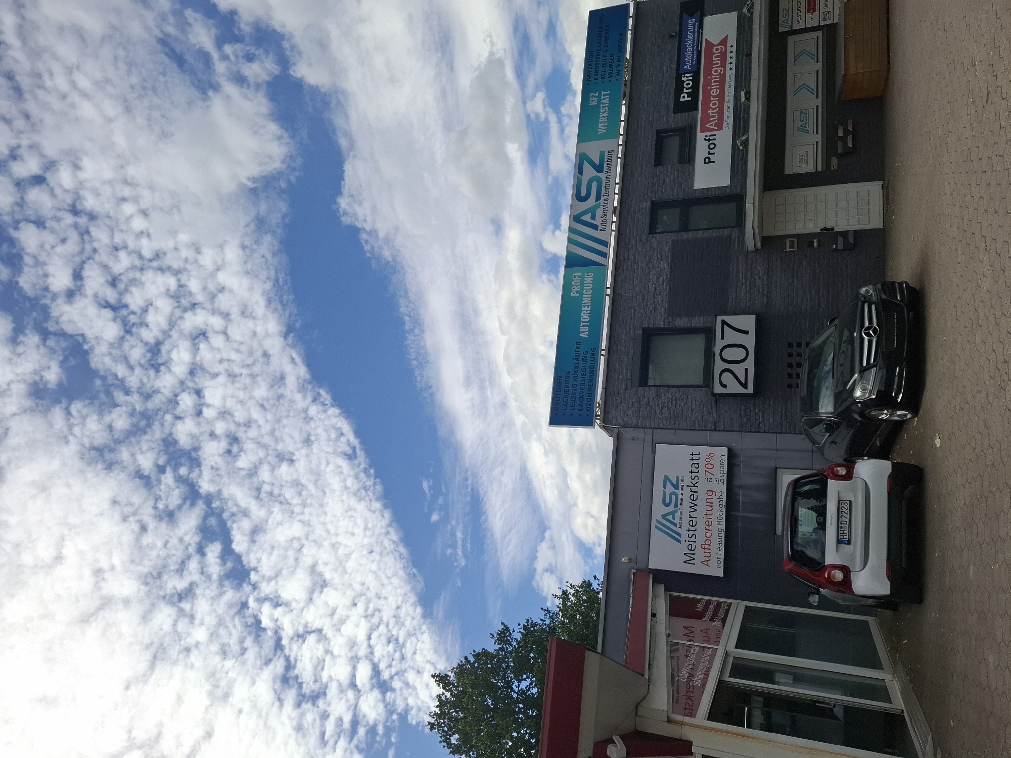 The image shows a building with several signs for automotive services in Hamburg, Germany. The building is dark gray brick and has a large sign that reads "ASZ Auto Service Zentrum Hamburg GmbH". Below this sign, there are signs for "Meisterwerkstatt" (master workshop), "Aufbereitung 70%" (reconditioning 70%), and "vor Leasing-Rückgabe" (before lease return). There are also signs for "Profi Autoreinigung" (professional car cleaning) and "Profi Autolackierung" (professional car painting).
Two cars are parked in front of the building: a white and red Smart car and a black Mercedes-Benz sedan. The sky is blue with scattered white clouds. The building appears to be a commercial establishment with a street-facing entrance and windows. The overall scene suggests a car service and repair shop.