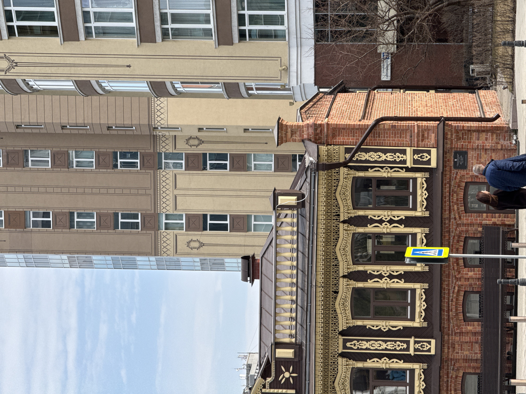 The image shows an architectural juxtaposition in Yekaterinburg, Russia, featuring a traditionally ornamented wooden house alongside a modern building. The wooden house exhibits ornate carvings and brick accents, and a blue and yellow pedestrian crossing sign is visible in front. Adjacent to it is a tall, multi-story building with a neutral color scheme and rectangular windows. The sky is partially visible above the buildings. Two people, a man and a woman, are standing by the wooden house, seemingly observing it.