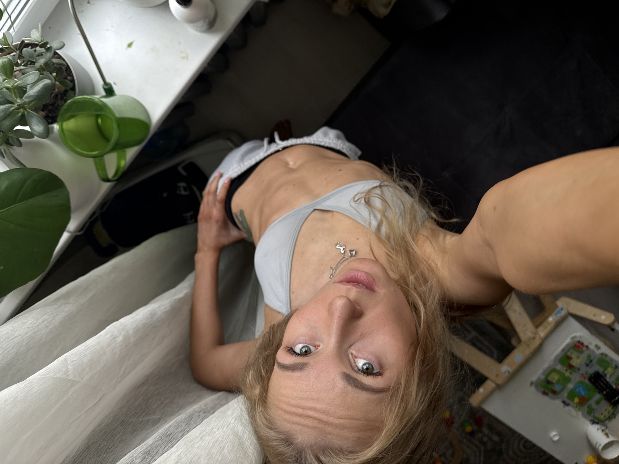 A fair-skinned woman with blonde hair is posing for a selfie. She is wearing a light gray sports bra and matching shorts. Her stomach is visible. She is also wearing a delicate necklace with small heart-shaped pendants. Her hand is resting on her hip. She has green eyes and appears to be looking directly into the camera.
The background includes a windowsill with several plants, including a succulent and a larger green plant. There is also a green watering can. A dark-colored couch is partially visible. An easel with a children's toy is located to the right of the woman. White curtains hang in the lower left corner of the image. The location of the photo is not indicated in the city of Chaikovsky, Russia.