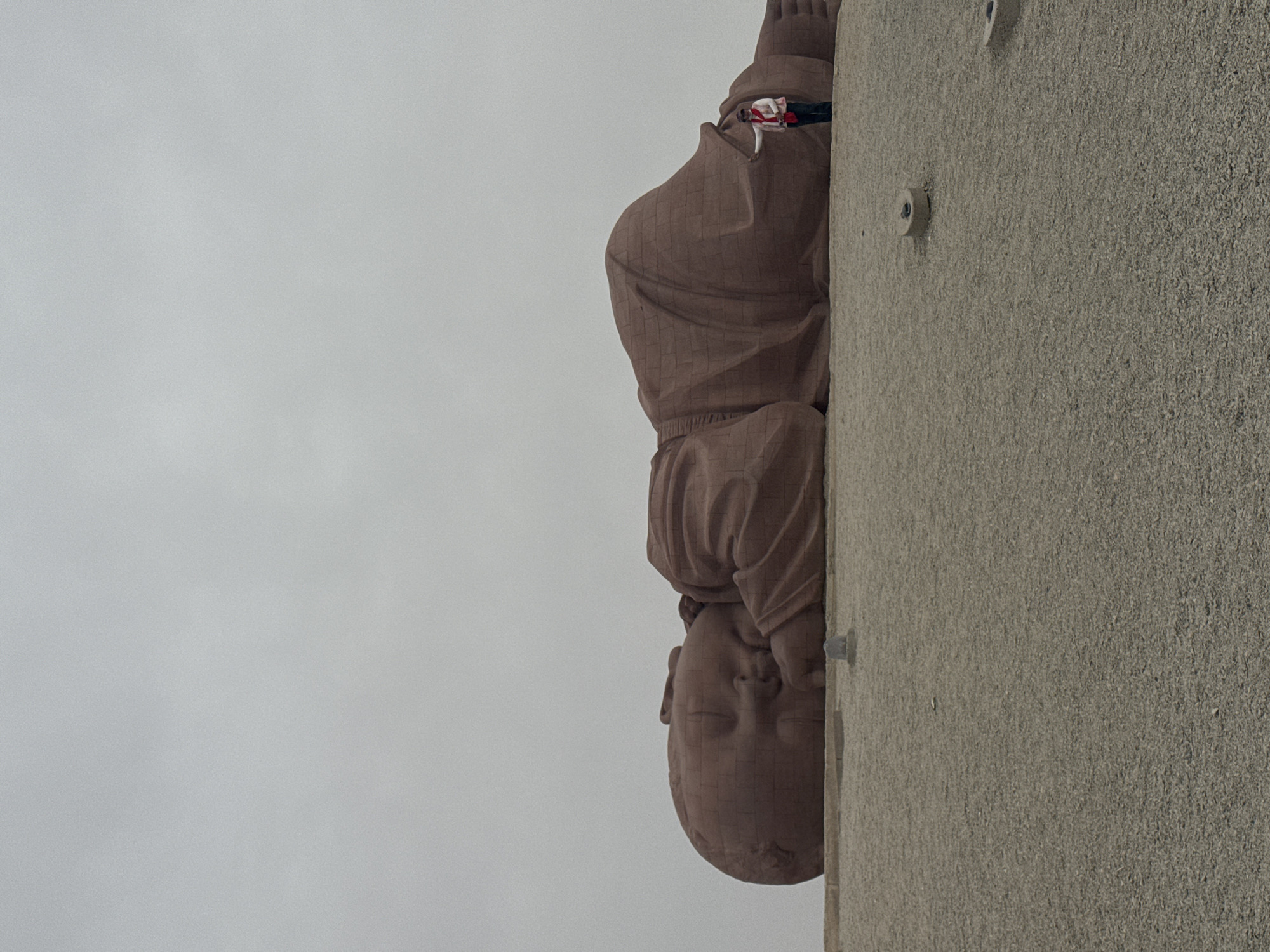 The image depicts a portion of the Statue of Xuanzang in Guazhou County, China, situated against a grey sky. The statue, made of brick or stone, shows the head and torso of a robed figure adjacent to a rough, concrete wall. The statue's gaze is directed downward, and it appears to be weathered. Fasteners are visible on the concrete wall.