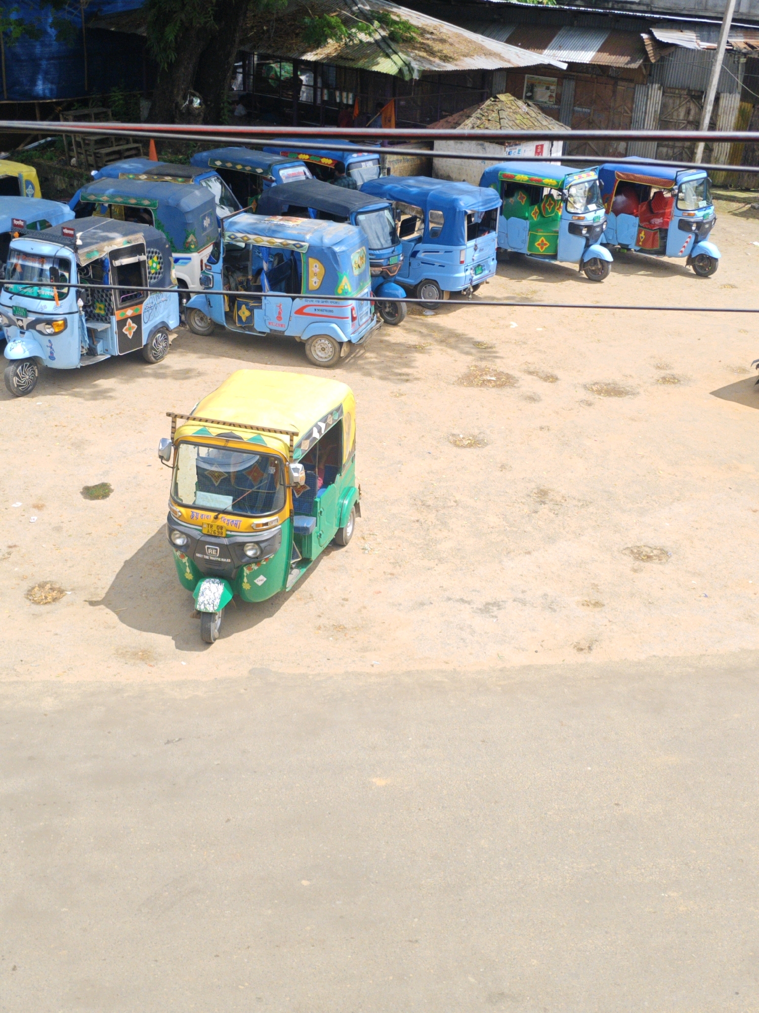 The image shows a street scene in Belonia, India, with several auto-rickshaws parked. In the foreground, a green and yellow auto-rickshaw is parked, facing left. It has a yellow roof and green body. The driver is partially visible through the windshield. Behind this auto-rickshaw, several other auto-rickshaws are parked in a row. These are mostly blue, with some yellow and red accents. They appear to be in a lot or on a wide street. In the background, there are some buildings with corrugated metal roofs, suggesting a commercial or informal settlement area. There are also some trees and power lines visible. The ground is dirt and gravel. The overall impression is a typical scene from a town or city in India, likely a transportation hub or parking area for these vehicles.