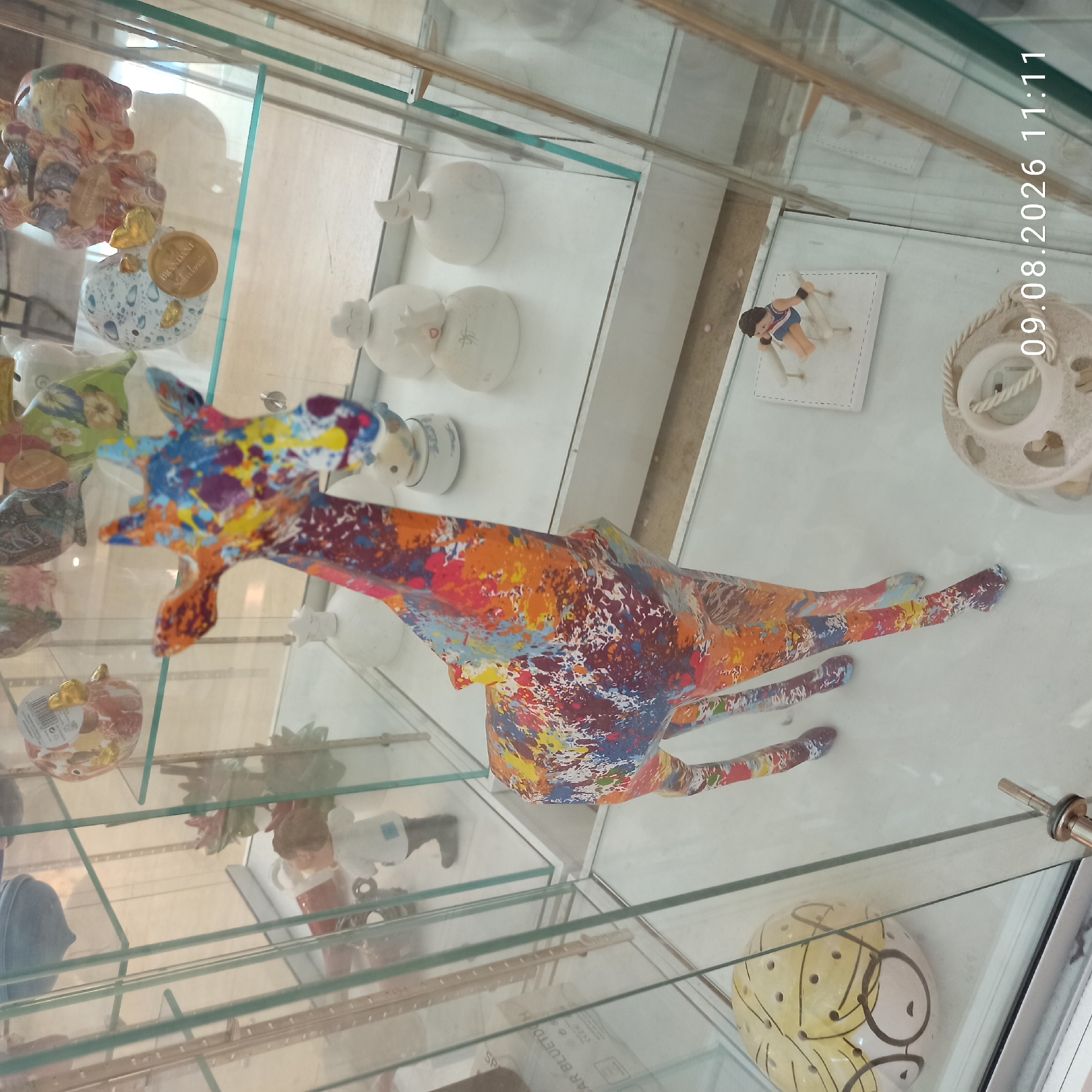 The image depicts a display cabinet filled with various decorative items, likely in a shop or home setting. In the center of the cabinet, a colorful, abstractly painted giraffe figurine stands prominently. The giraffe is made of ceramic or a similar material and features a vibrant, splattered design of blues, oranges, purples, and yellows.
To the left of the giraffe, there is a large, spherical object decorated with a cartoonish face, resembling a yellow head with glasses and dotted patterns. Further to the left, partially visible, are other ceramic objects, including what appears to be a gnome-like figurine.
To the right of the giraffe, there are several small, white ceramic figurines. One is round with a crescent moon on top, another has a crown shape, and others are spherical with subtle decorative elements. In the background, various other decorative pieces are visible, including bird-shaped piggy banks with intricate patterns.
The display cabinet itself is made of glass and metal, with visible shelves and vertical supports. The lighting in the scene is diffused, creating reflections on the glass surfaces. In the lower right corner, a timestamp reads "09.08.2026 11:11", indicating the date and time the photograph was taken.
This scene does not provide any specific details or context to place it within the city of Molfetta, Italy. The image focuses solely on the contents of the display cabinet.