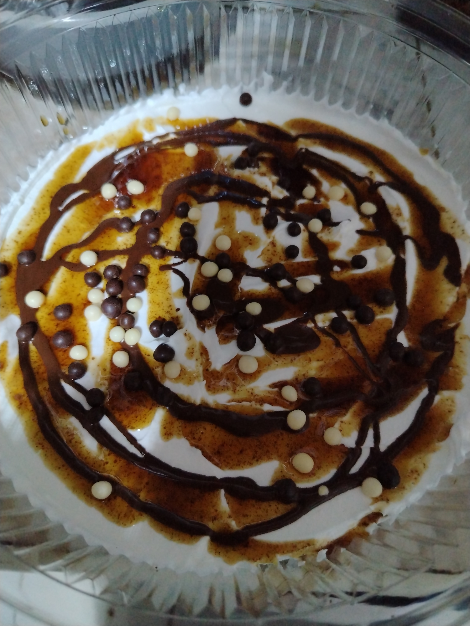 The image shows a dessert inside a clear plastic container. The dessert has a white base, likely whipped cream, and is decorated with a concentric pattern of brown sauce, dark chocolate drizzle, and scattered white and dark chocolate balls. The photograph was taken indoors in Tinaquillo, Venezuela.