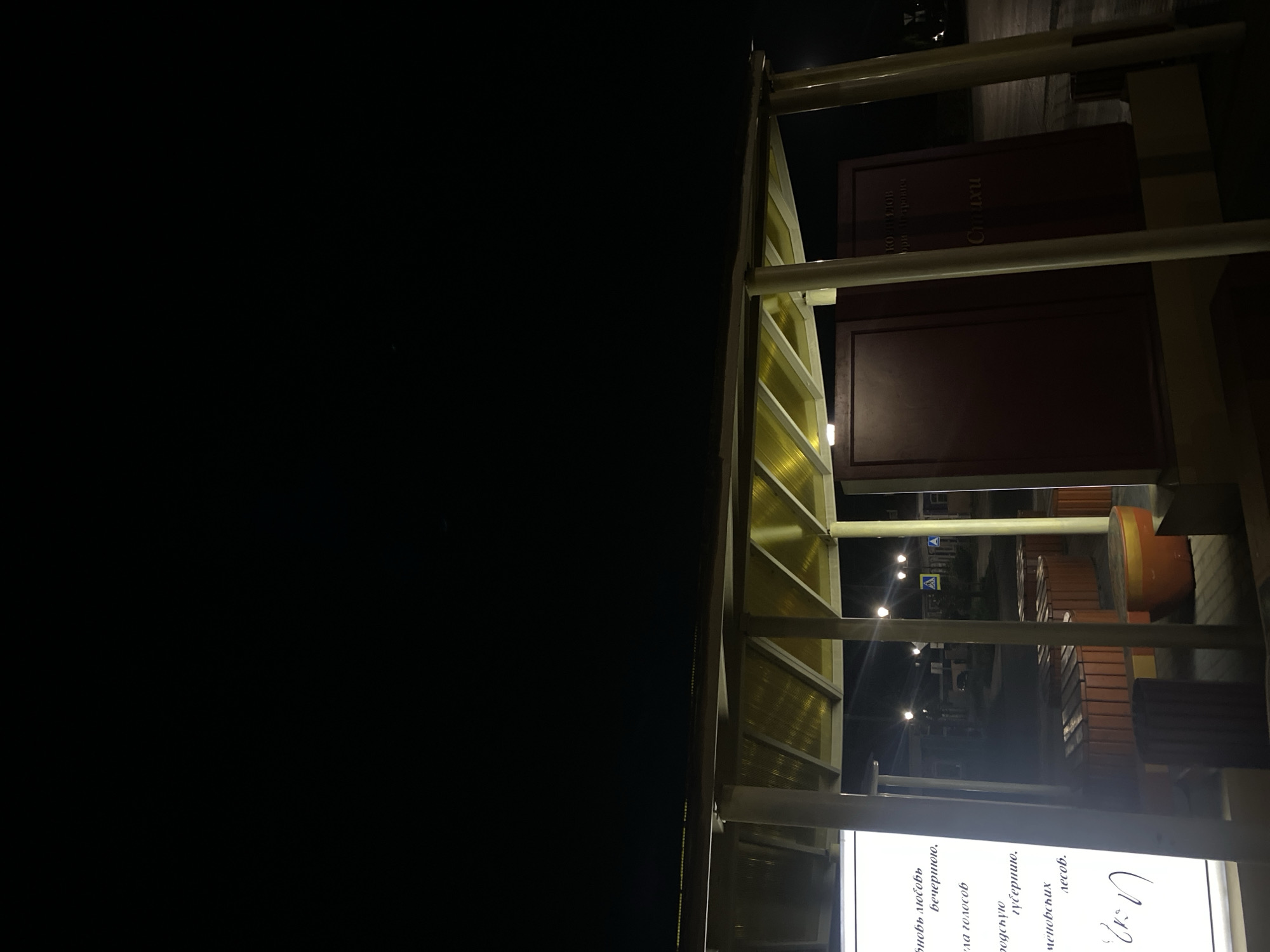 The image, captured at night, presents an illuminated outdoor public space. In the foreground, a contemporary architectural structure, possibly a canopy or shelter, is prominent. It features a light-colored, possibly metallic, framework supporting a translucent, corrugated yellow roof that emits a warm glow from internal or nearby lighting.
Behind this structure, to the right, two dark rectangular signs are mounted, one above the other. OCR identifies Russian text on these signs: "Кори Петрович" (Kory Petrovich) and "Стихи" (Poems/Verses), suggesting a tribute or informational display for a poet. Further back, several circular, multi-tiered benches with wooden slats and yellow-orange accents are visible, alongside a dark-colored trash bin.
To the bottom left, partially covered by the canopy, a large, brightly lit white sign displays more Russian text, likely poetic excerpts: "Вновь любовь" (Again love), "Вечернюю," (Evening,), "ела голосов" (ate voices), "одскую" (city/urban), "губернию," (province/governorate), "мёновских" (of Menov), "лесов." (forests.). A distinctive signature-like graphic is also present on this sign.
In the background, multiple streetlights cast white light, and a blue-and-white pedestrian crossing sign can be faintly seen, indicating a road or path. The scene is devoid of people, depicting a quiet, artificially lit urban or park setting after dark under clear weather conditions.
