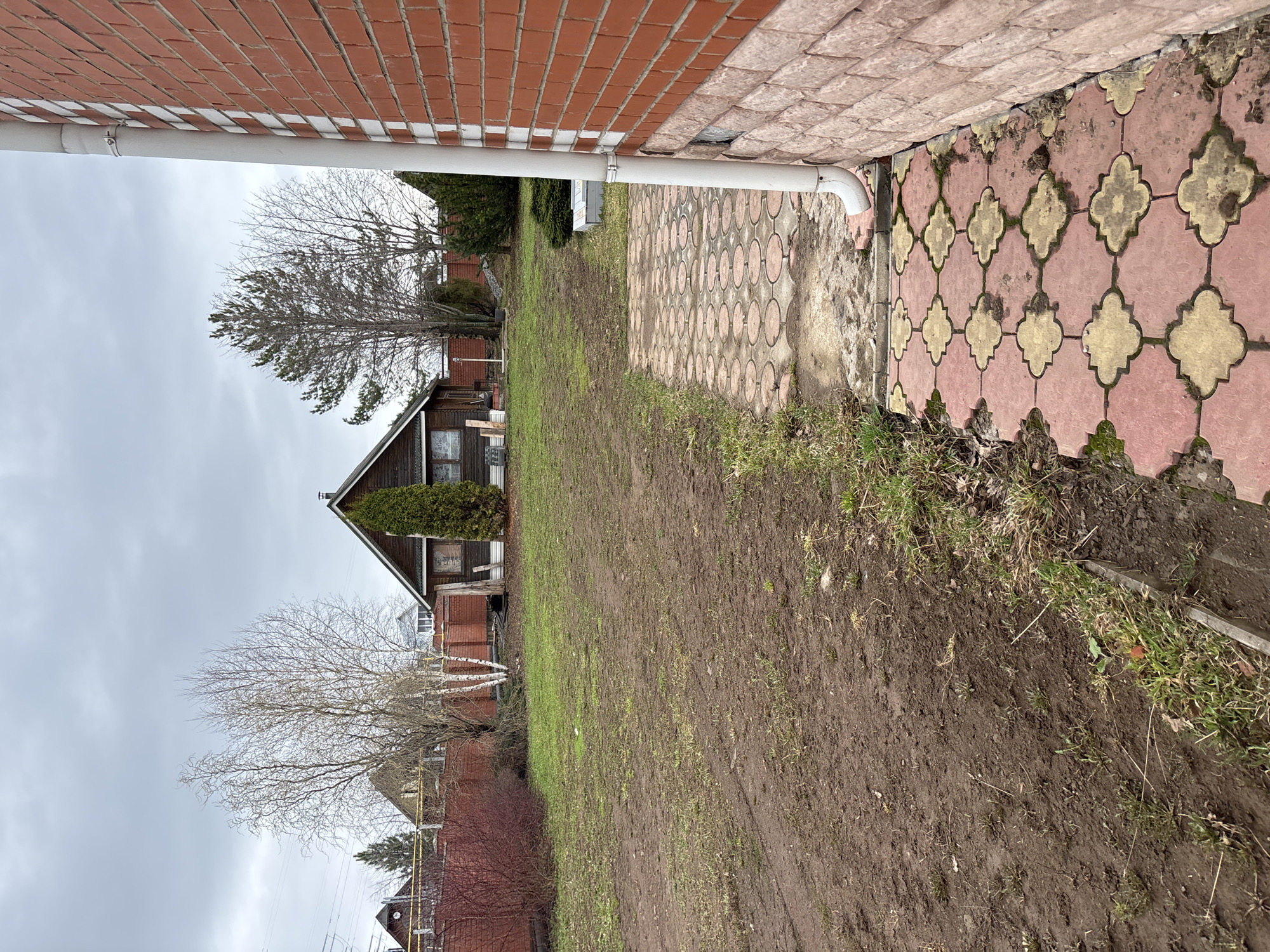 The image depicts a view of a yard in Ферма, Russia, on an overcast day. The foreground consists of muddy soil with sparse grass, suggesting recent wet conditions. To the right, there's a section of patterned paving stones, exhibiting signs of wear and moss growth. A white downspout runs along the edge of a brick building, indicating drainage infrastructure.
In the middle ground, a small, rustic wooden house with an A-frame roof is visible, flanked by bare trees and evergreen shrubs. The house appears to have at least one window and is partially obscured by vegetation.
The background shows neighboring buildings and more bare trees under a grey, overcast sky, giving the impression of a rural or suburban residential area. There are no people visible in the image. The overall scene conveys a sense of a quiet, somewhat neglected outdoor space.