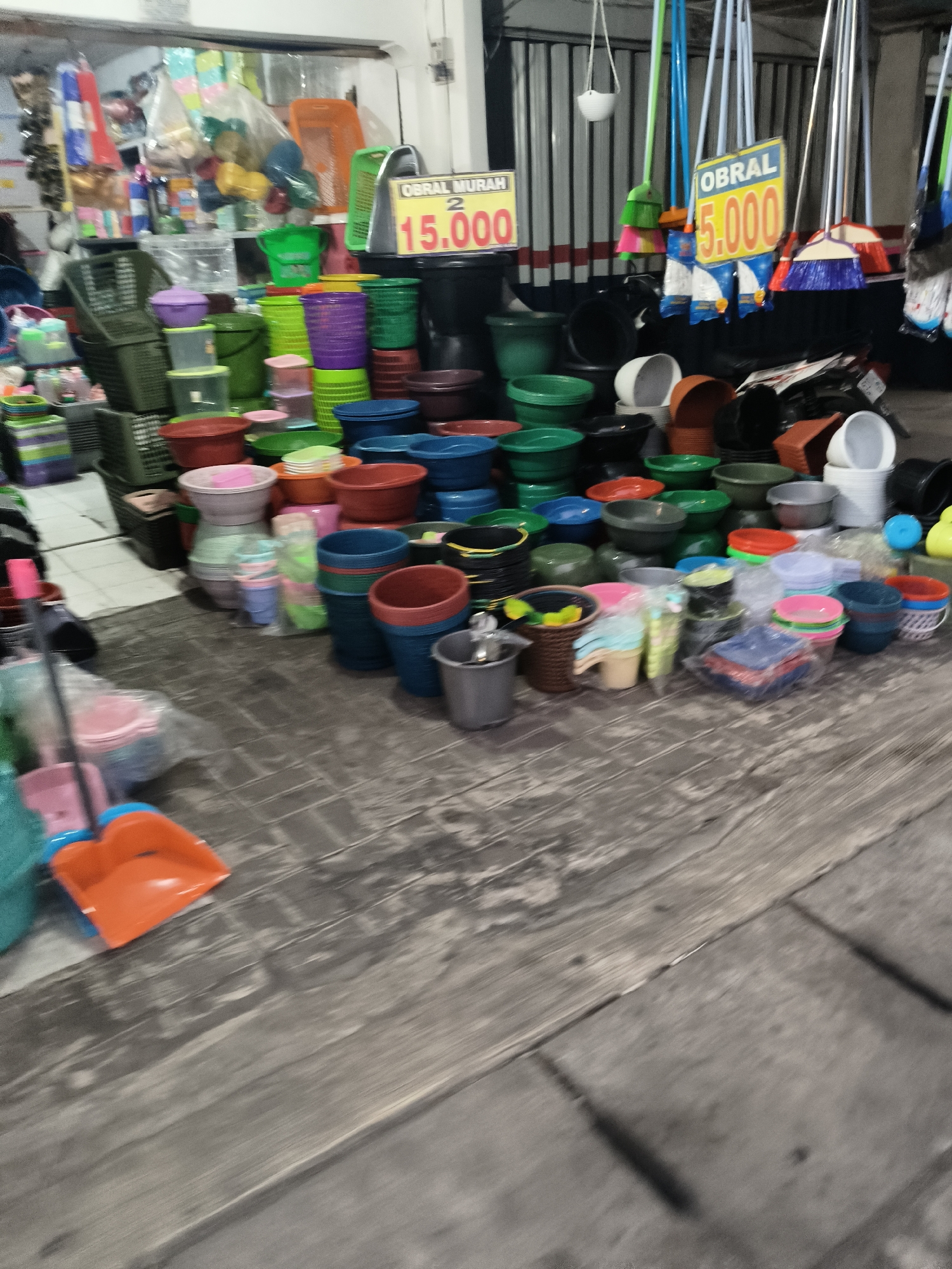 The image depicts a marketplace stall in South Tangerang, Indonesia, brimming with various plastic household items. The stall displays a colorful assortment of buckets, basins, and storage containers in a range of sizes and hues, including green, purple, blue, red, and orange. Among the items are brooms with bright handles, suggesting a general store or a stall specializing in cleaning supplies and kitchenware. Two signs are visible, one in yellow reading "OBRAL MURAH 2 15.000" and another in yellow and blue reading "OBRAL 5.000", indicating discounted prices, likely in Indonesian Rupiah. The stall is set up on a wooden surface, possibly a table or floor, and the background shows more stalls and what appears to be a covered market area. The lighting suggests it might be evening or an indoor market with artificial lighting.