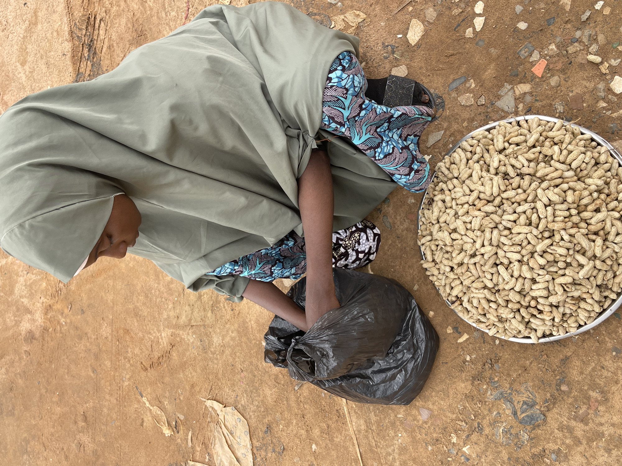 In Abuja, Nigeria, a young person wearing a light olive-green hijab and a blue and pink patterned dashiki is squatting on reddish-brown dirt. They are reaching into a large, black plastic bag with their left hand. In front of them, a shallow, circular metal tray is filled with peanuts in their shells. The ground around them is littered with scattered dry leaves and small stones.