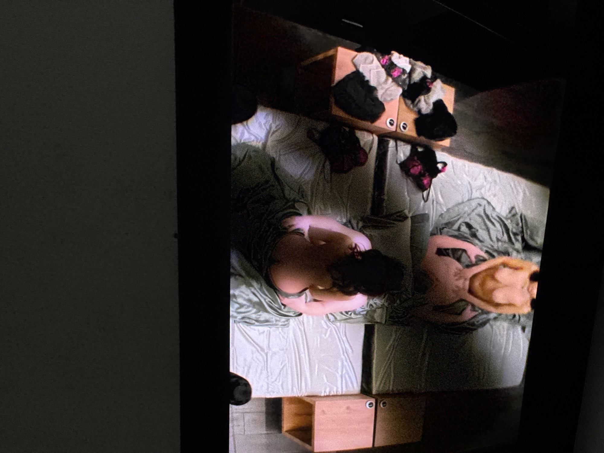 An overhead, full shot captures two individuals on a bed in an indoor setting. The person on the left is seen from the back, with their head down, appearing to be lying on their side under a light blue sheet. To the left of this individual is a wooden box, likely a storage container, on top of a dark gray surface. The second person, on the right, is lying on their back, facing the camera, with their head and upper torso exposed. They are partially covered by the same light blue sheet. Beside them, to the right, is another wooden storage unit. This unit is piled with various articles of clothing, predominantly undergarments in shades of black, white, and pink. The scene is dimly lit, casting shadows across the bed and surrounding objects.