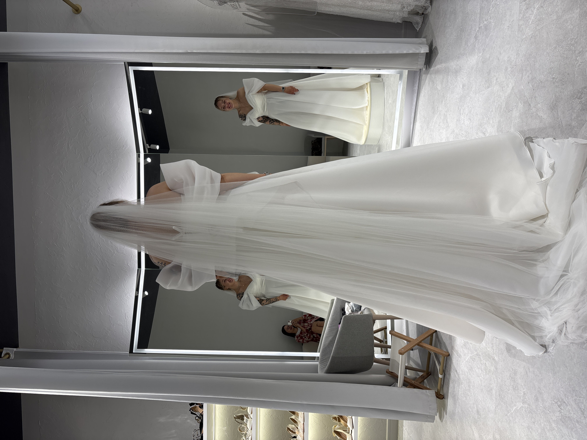 The image depicts a bridal shop fitting room in Krasnoyarsk, Russia. The primary subject is a person wearing a white wedding dress and veil, viewed from behind, with their reflection visible in a large, illuminated mirror. The reflection shows the person smiling, with visible tattoos on their arms. In the background, a second person is seated, holding a phone, and appears to be taking a picture. To the left of the main subject, a rack of shoes is visible on shelves. The room has a modern, minimalist aesthetic with grey flooring and white curtains.