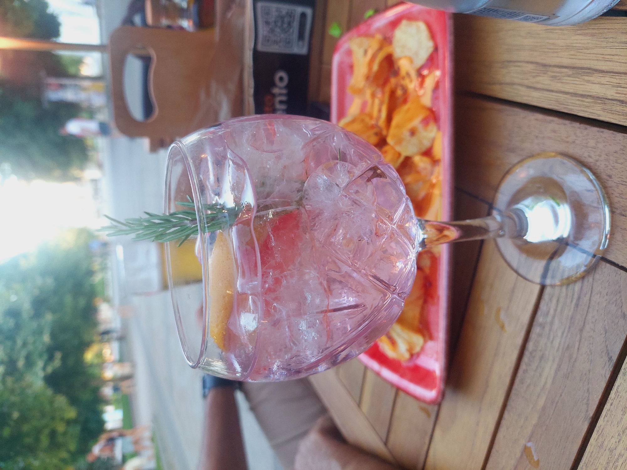 The image shows a close-up of a pink gin and tonic cocktail in a stemmed glass on a wooden table. The drink contains ice, a slice of citrus, and a sprig of rosemary. In the background, out of focus, is a red plate of potato chips, a brown paper bag with text that appears to say "ento" and a QR code, a blurry street scene with trees and people walking, and a glass of beer. The lighting suggests it is daytime, possibly late afternoon, with sunlight filtering through the trees.