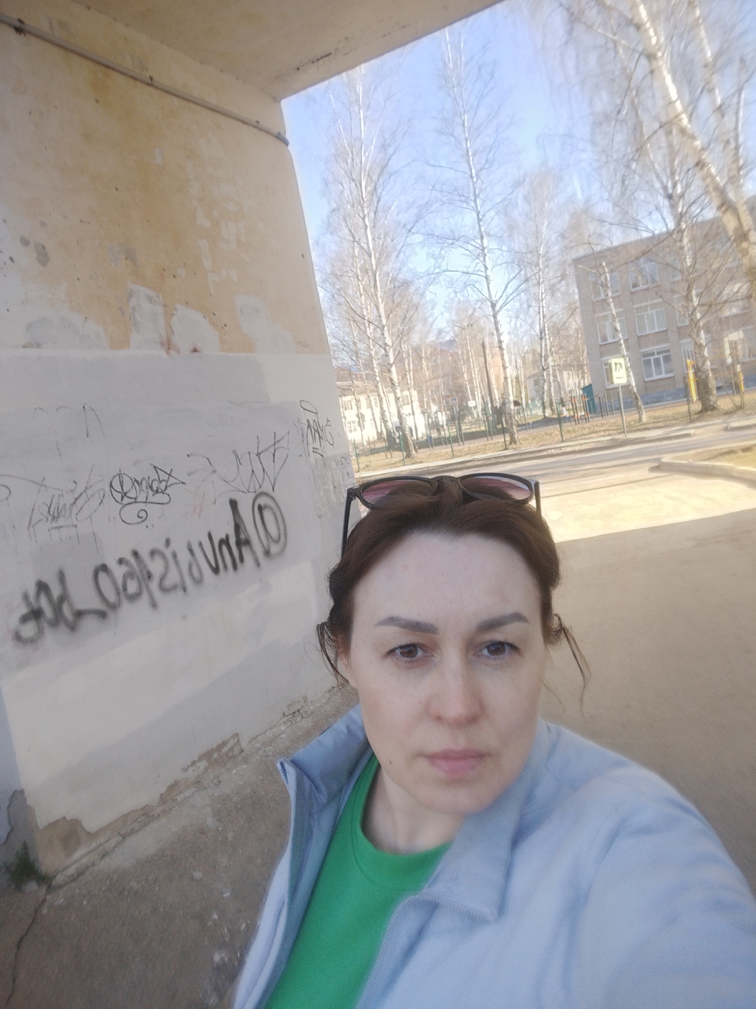 A woman is captured in a selfie, looking directly at the camera with a neutral expression. She has brown hair, dark eyebrows, and wears sunglasses pushed up onto her head. She is dressed in a green t-shirt under a light blue jacket.
To the left of the woman is a weathered, light-colored building wall, possibly part of an underpass or an old structure. The wall is adorned with black, stylized graffiti, which appears mirrored, displaying shapes and letters that resemble "DnA" along with other illegible scrawls. The wall itself shows signs of age, with peeling paint and discoloration.
The background reveals a clear, bright blue sky, indicating a sunny day. Numerous bare deciduous trees, possibly birches, stand against the sky, suggesting late winter or early spring. To the right, a paved road or path curves into the distance. Further back, a multi-story building with many windows, characteristic of urban architecture, is visible. A small sign, possibly indicating a pedestrian crossing, can be seen near the road. The overall scene depicts a casual outdoor moment in an urban environment, consistent with a setting like Chaikovsky, Russia, during the colder months.