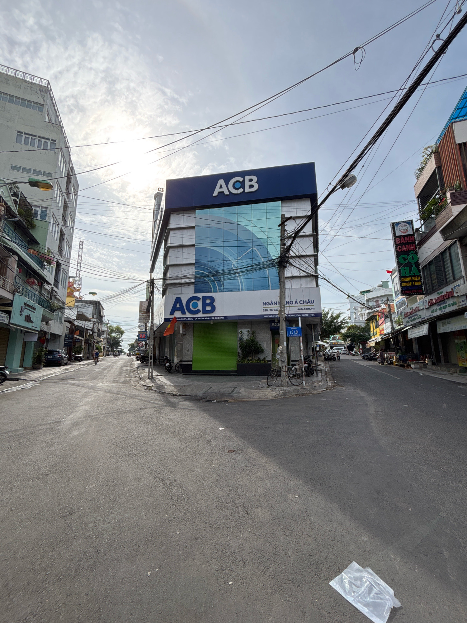 The image depicts a street scene in Nha Trang, Vietnam. A modern bank building with the logo "ACB" is prominently featured in the foreground, with its blue glass facade reflecting the sky. To the left of the ACB building, a multi-story building with a light green exterior is visible, followed by other shops and buildings lining the street. The street itself is paved with asphalt and appears to be a typical urban road with various vehicles, including motorcycles and a car, parked along the sides and in motion. A person is seen riding a bicycle in the distance. Overhead, a complex network of electrical wires crisscrosses the sky, typical of many Southeast Asian cities. The sky is partially cloudy, with the sun creating some glare. Several shops with Vietnamese signage are visible on the right side of the street. The overall impression is a bustling urban environment during daylight hours.