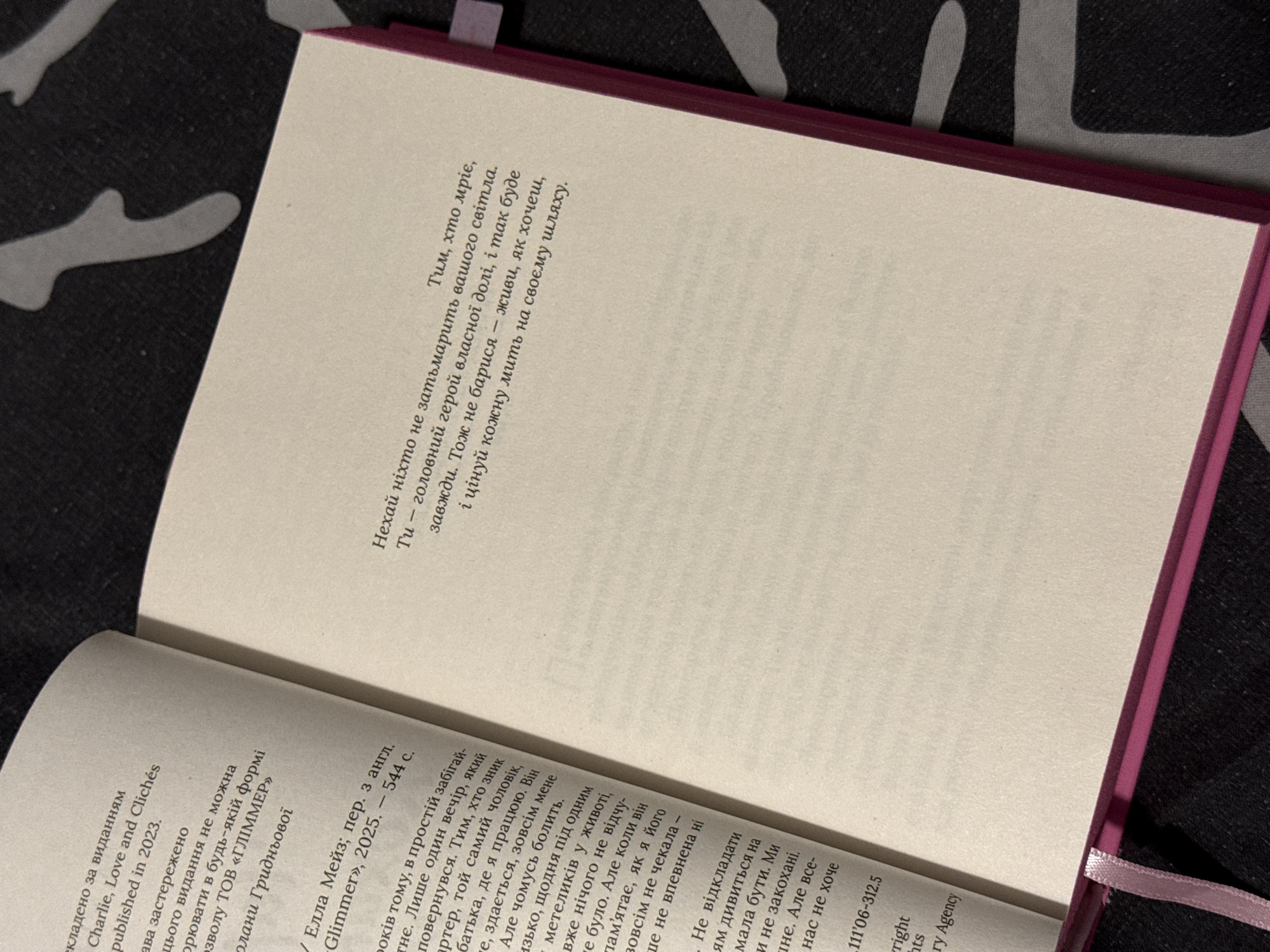 An open book with pages of text is laid flat on a dark, patterned surface. The book is bound in pink and has a pink ribbon bookmark peeking out from between the pages. The text is in Ukrainian. The left page contains information about the publication, including the title "Charlie, Love and Clichés" and the publication year "2023". The right page contains a quote in a slightly larger font: "Ніхто не затьмарить вашого світла. Тим, хто мріє, Ту — головний герой власної долі, і так буде завжди. Тож не барися — живи, як хочеш, і цінуй кожну мить на своєму шляху." This translates to: "No one will overshadow your light. He who dreams, You - the main character of your own destiny, and so it will be always. So don't delay - live as you wish, and cherish every moment on your path." The image does not contain any information that would allow for the determination of its location in the city of Łódź, Poland.