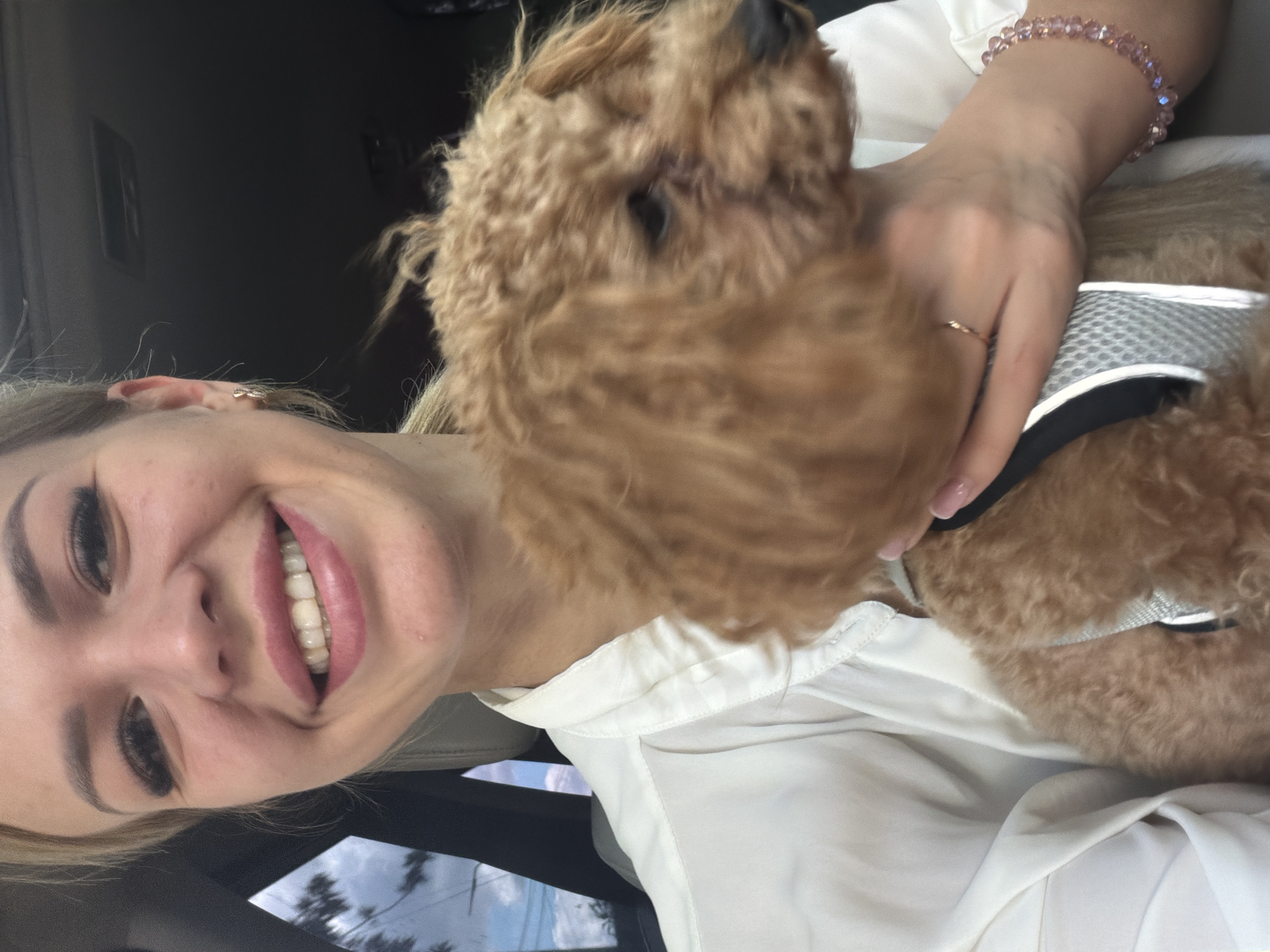 A young Caucasian woman with blonde hair and long eyelashes is holding a small, fluffy, light brown poodle in the passenger seat of a car. She is smiling broadly, and her teeth are visible. The woman is wearing a white collared shirt and a delicate gold ring on her left hand. The poodle is wearing a grey harness. The background of the image shows the interior of a car, with a cloudy sky and trees visible through the window. There is no indication in the image that this scene is located in Kyiv, Ukraine.
