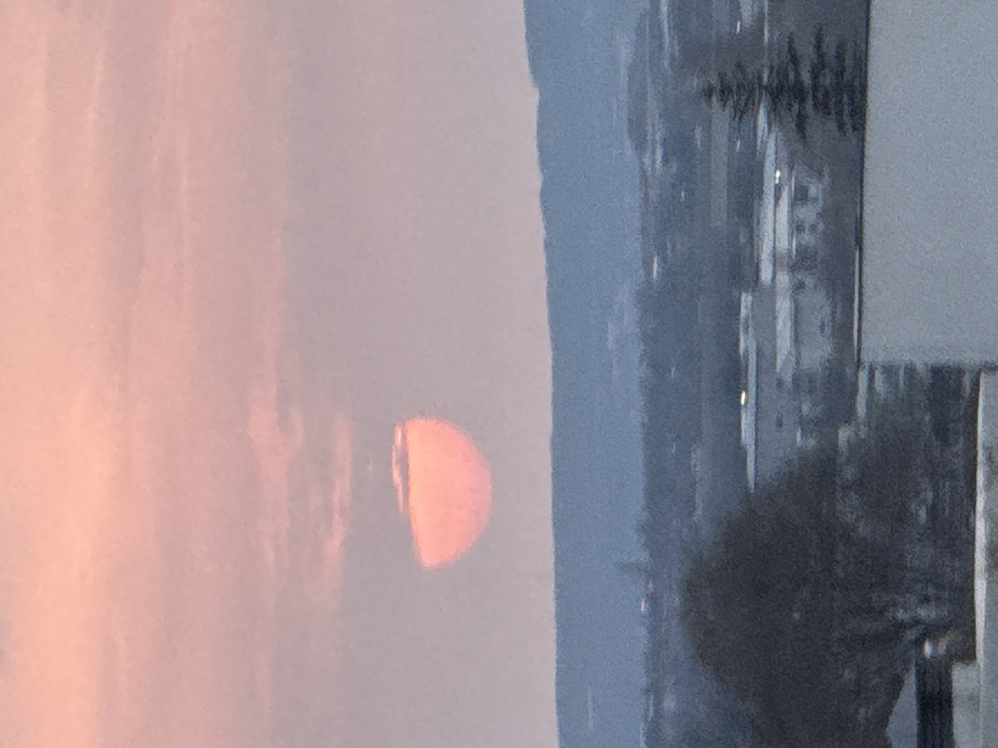 A horizontal view shows a sunrise or sunset next to a hazy cityscape. A large, half-disk of orange sunlight sits left of center, over a layer of blue, hazy land. The left side of the image shows a pale pink sky with subtle vertical streaks. The right side of the image depicts a blurred cityscape, with buildings and trees visible. It is daytime. The photo was most likely taken in Varna, Bulgaria.