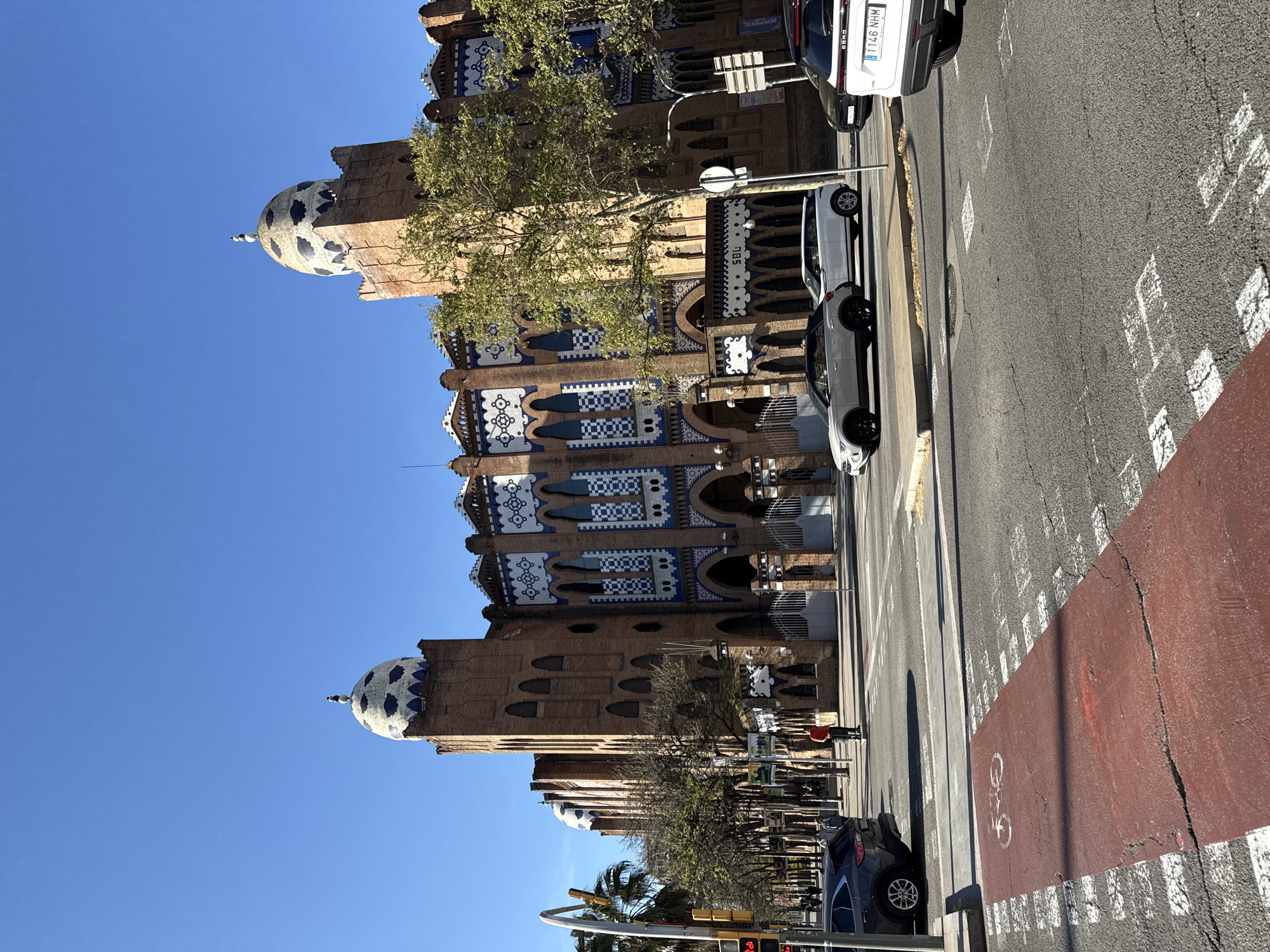 The image depicts the exterior of the Monumental bullring in Barcelona, Spain. The building has a distinct neo-Mudéjar architectural style with brickwork, arched windows, and decorative blue and white tiles. Two domed towers are visible. The bullring fronts a street with cars and a bike lane. A few trees are present. The sky is clear and blue.