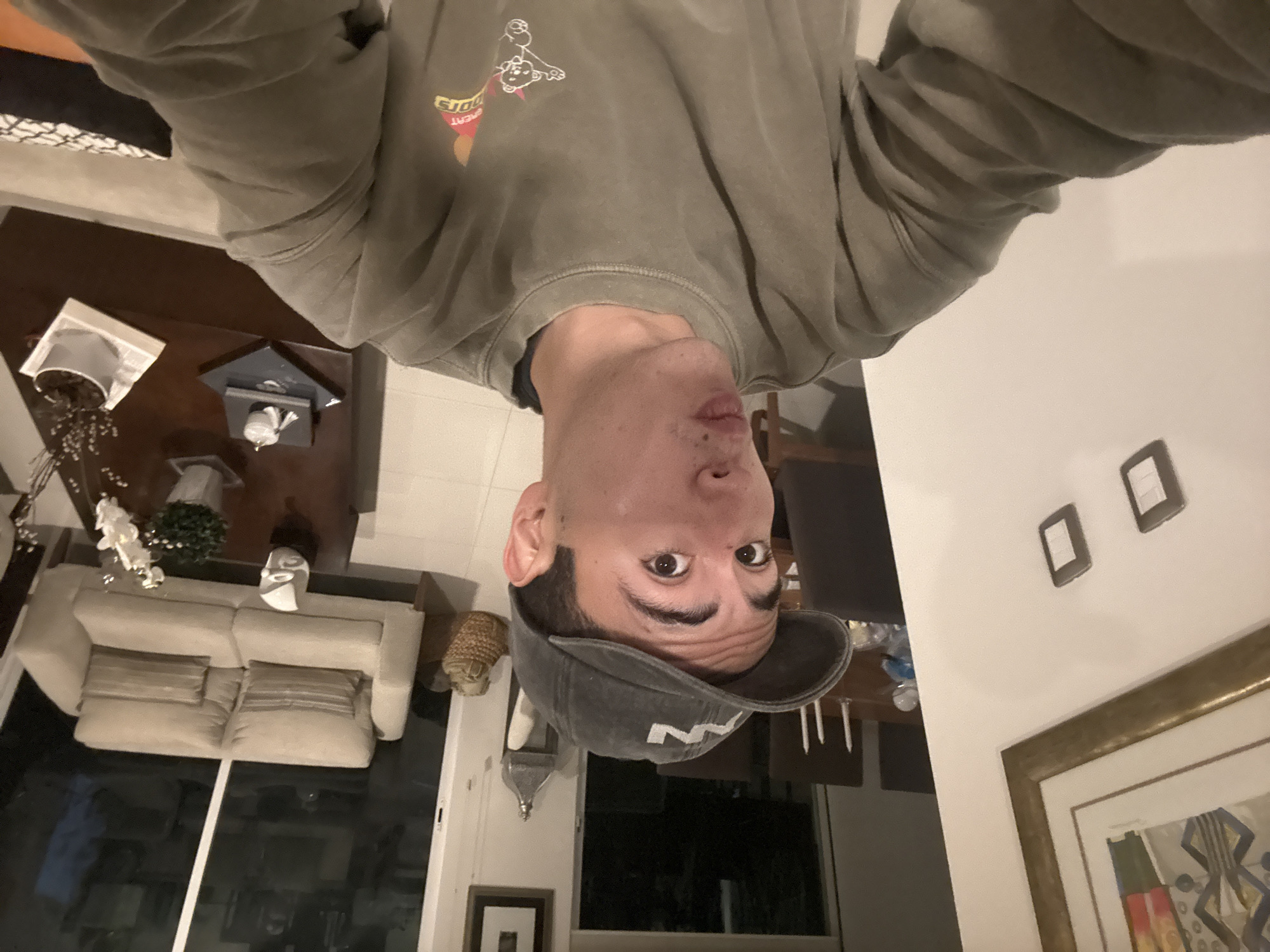 A photo taken by a person wearing a gray sweatshirt and a baseball cap with a white letter "M" on it, is upside down. The person is looking directly at the camera. In the background, there is a living room with a white couch, a wooden table, and a painting on the wall. Based on location data, it appears the photo was taken in San Bernardino Tlaxcalancingo, Mexico.